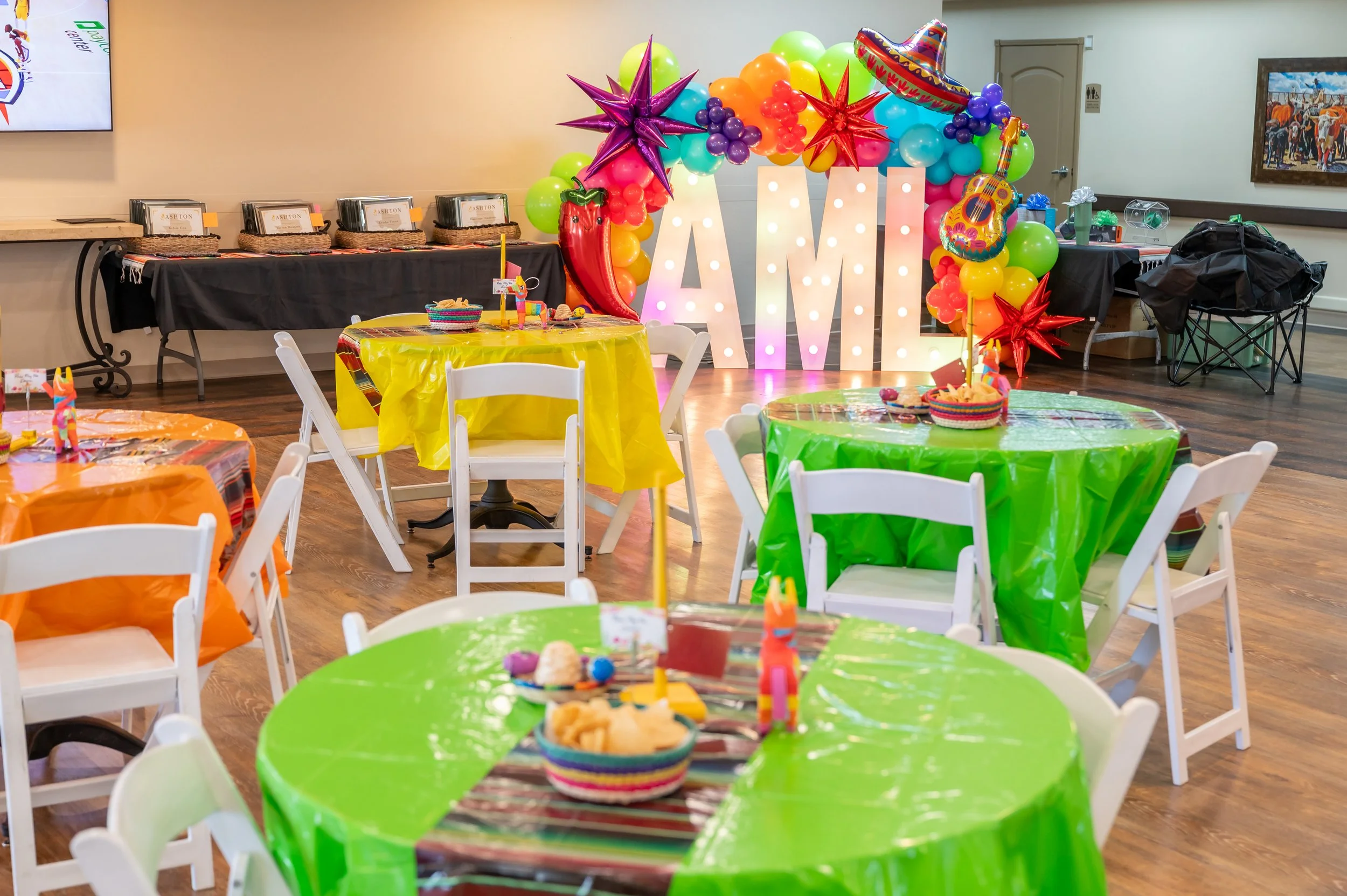 Colorful fiesta-themed birthday party setup with green, orange, and yellow tables, balloons, a large illuminated 'AMi' sign, and a decorated backdrop with sombrero and guitar.