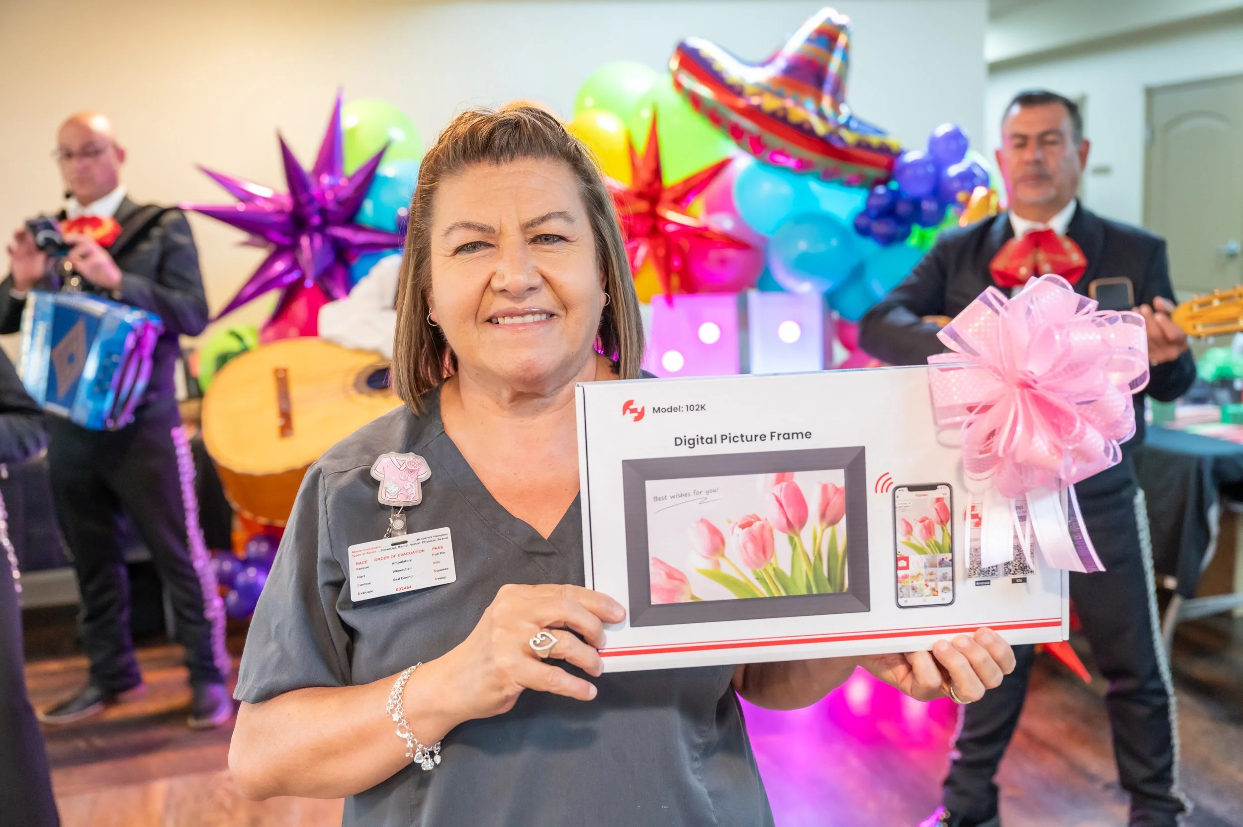 A woman smiling and holding a boxed digital picture frame at a festive celebration with balloons and balloons in the background, accompanied by musicians.