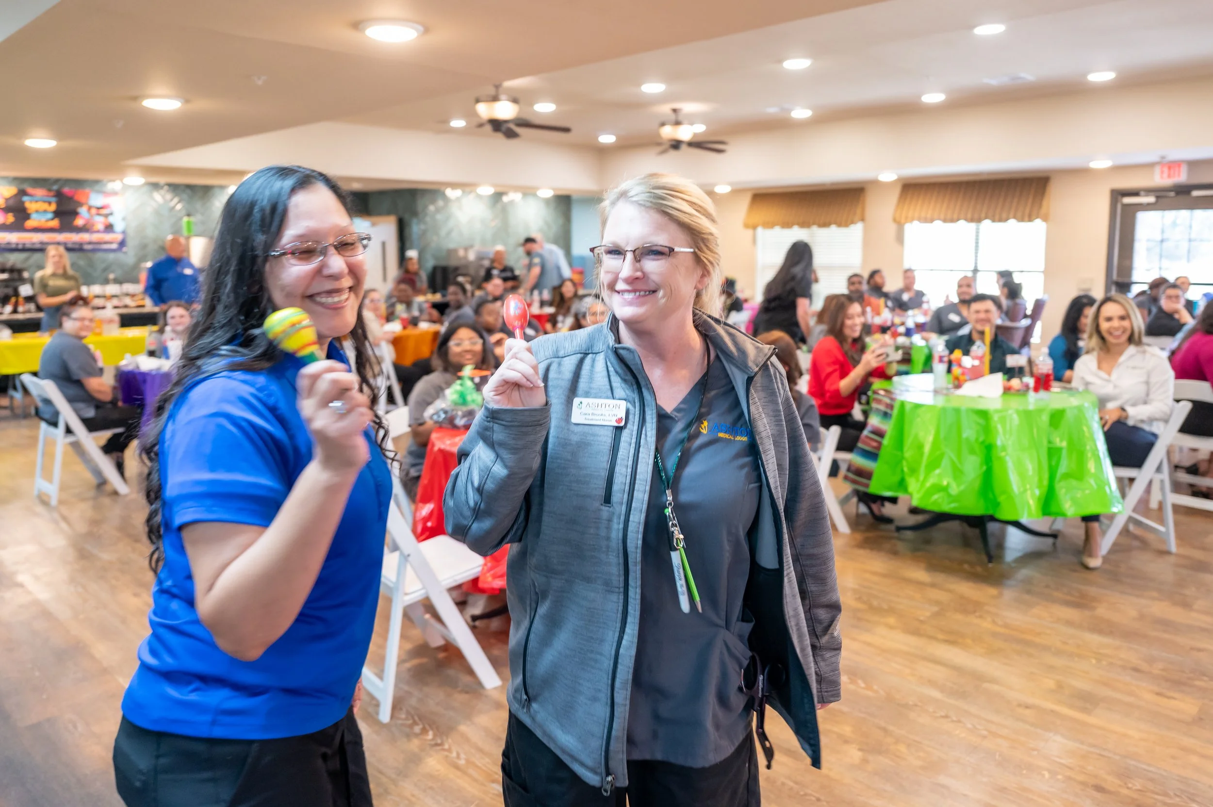 Two women smiling and holding small toys at a party or gathering with multiple people seated at colorful tables in a large, well-lit room.