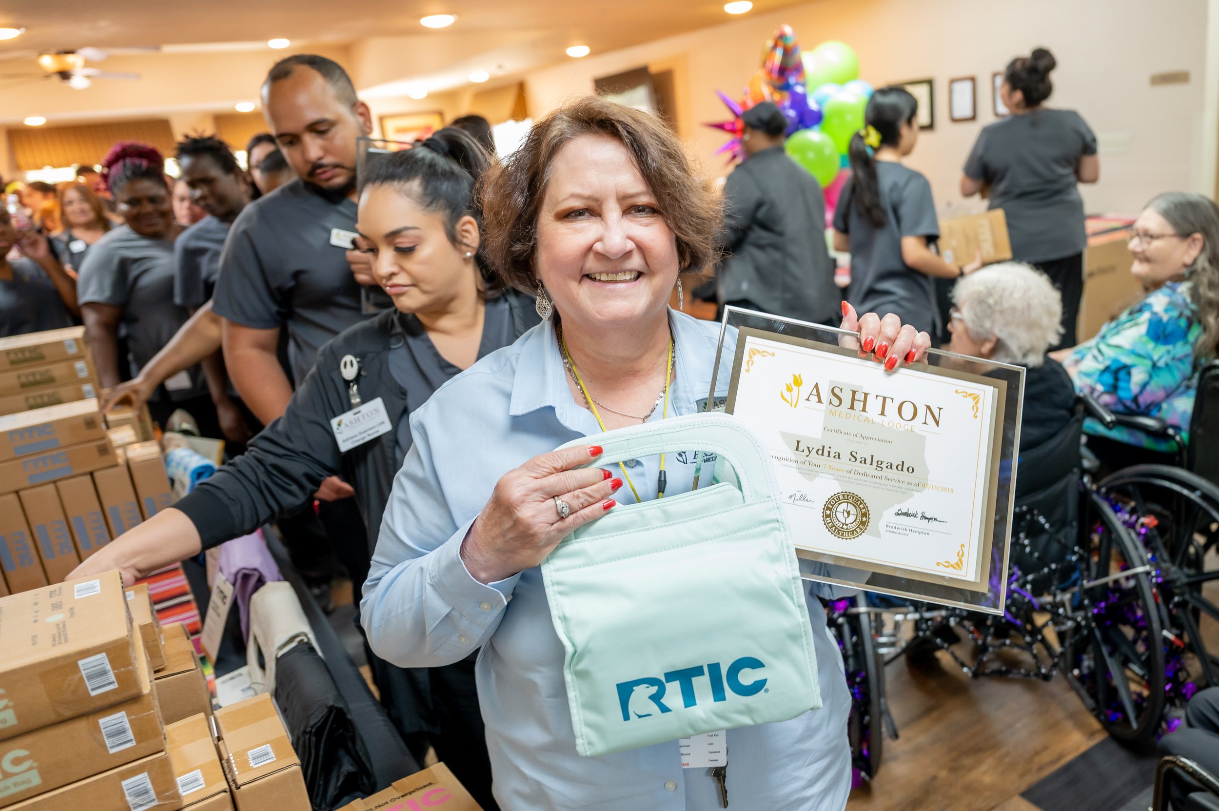 A woman smiling and holding a certificate of appreciation and a white tote bag with the RTIC logo at a community event. She is surrounded by people, some in line with boxes and others in wheelchairs, in a decorated indoor space.