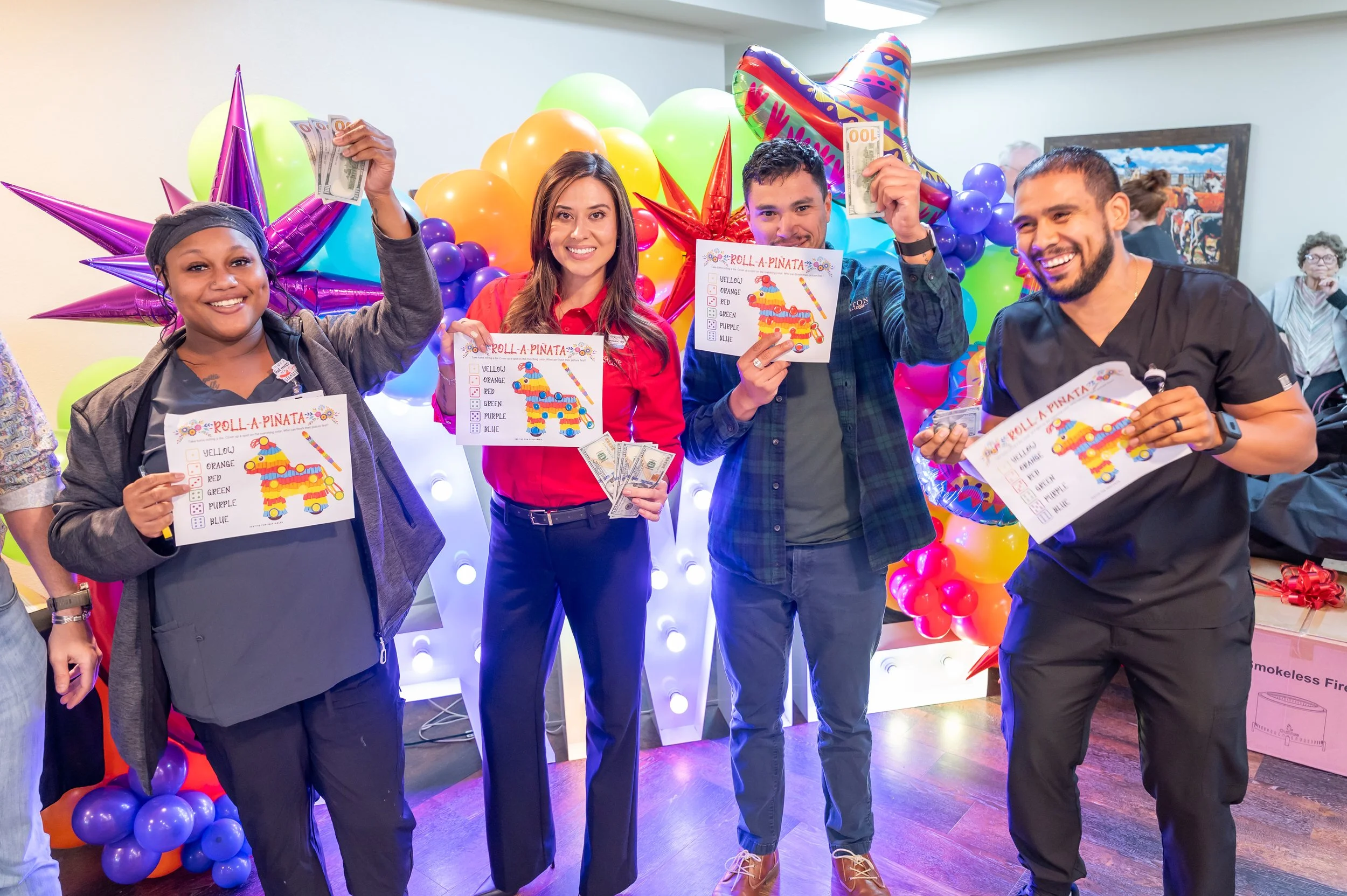 Group of four people celebrating a birthday party, holding envelopes and small cash, standing in front of colorful balloons and decorations, smiling at the camera.