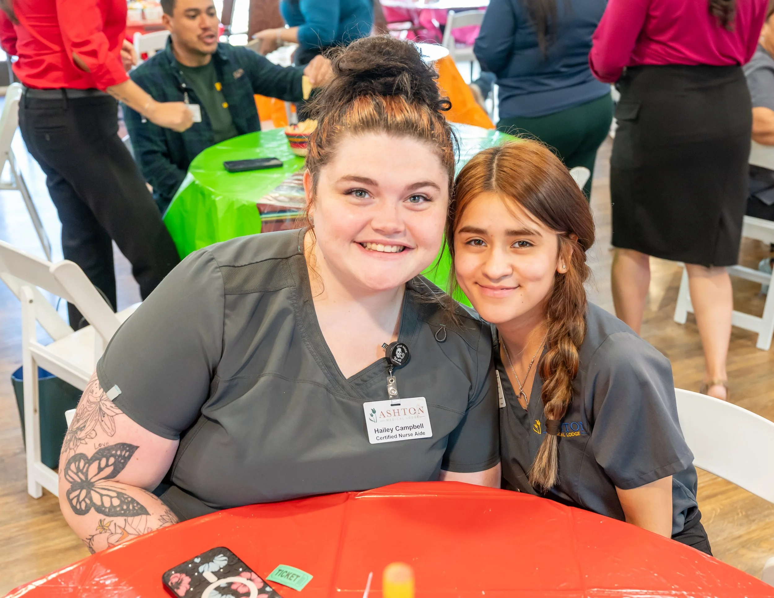 Two women smiling and sitting close together at a table during a social event. Behind them, other people are standing and talking, with some wearing colorful clothing and gathered around tables with bright tablecloths.