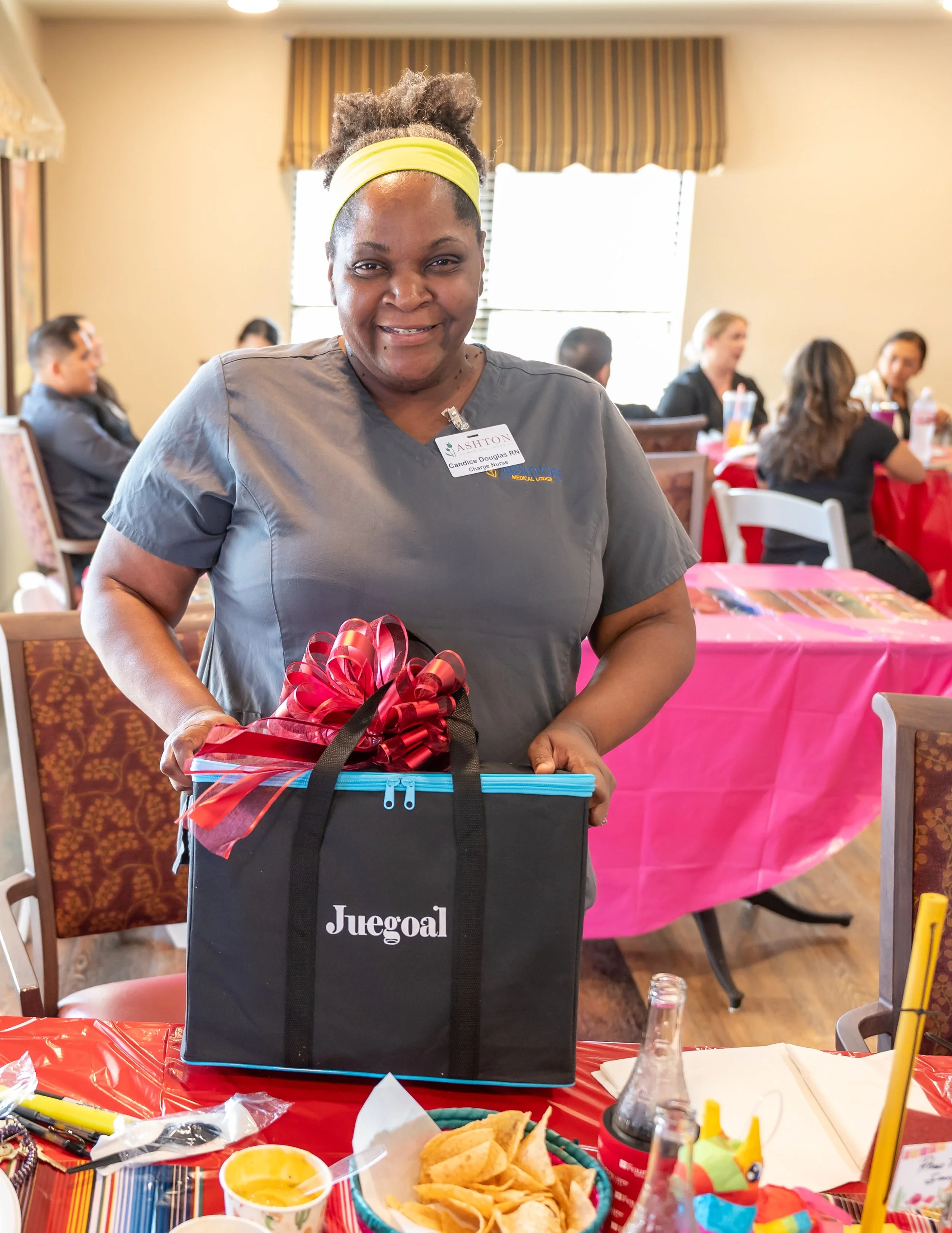 A woman with a yellow headband and gray medical scrubs standing at a decorated table, holding a gift bag with a red bow. She is smiling and wearing a name badge that reads 'Candice Douglas RN.' In the background, people are seated at tables with pink