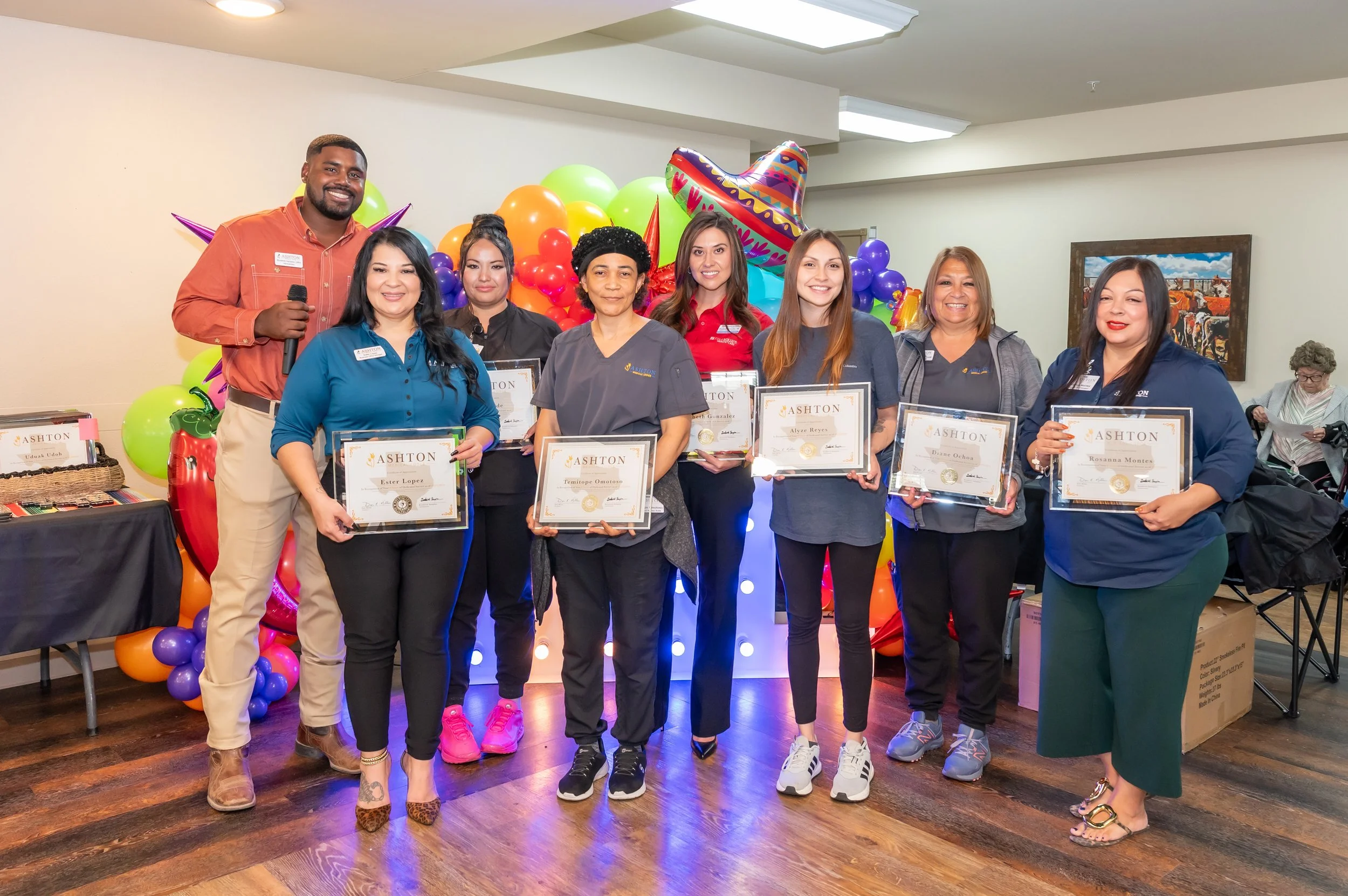 Group of eight people, six women and two men, standing indoors with colorful balloons and decorations in the background. They are holding certificates, smiling, dressed casually for an award or recognition event.