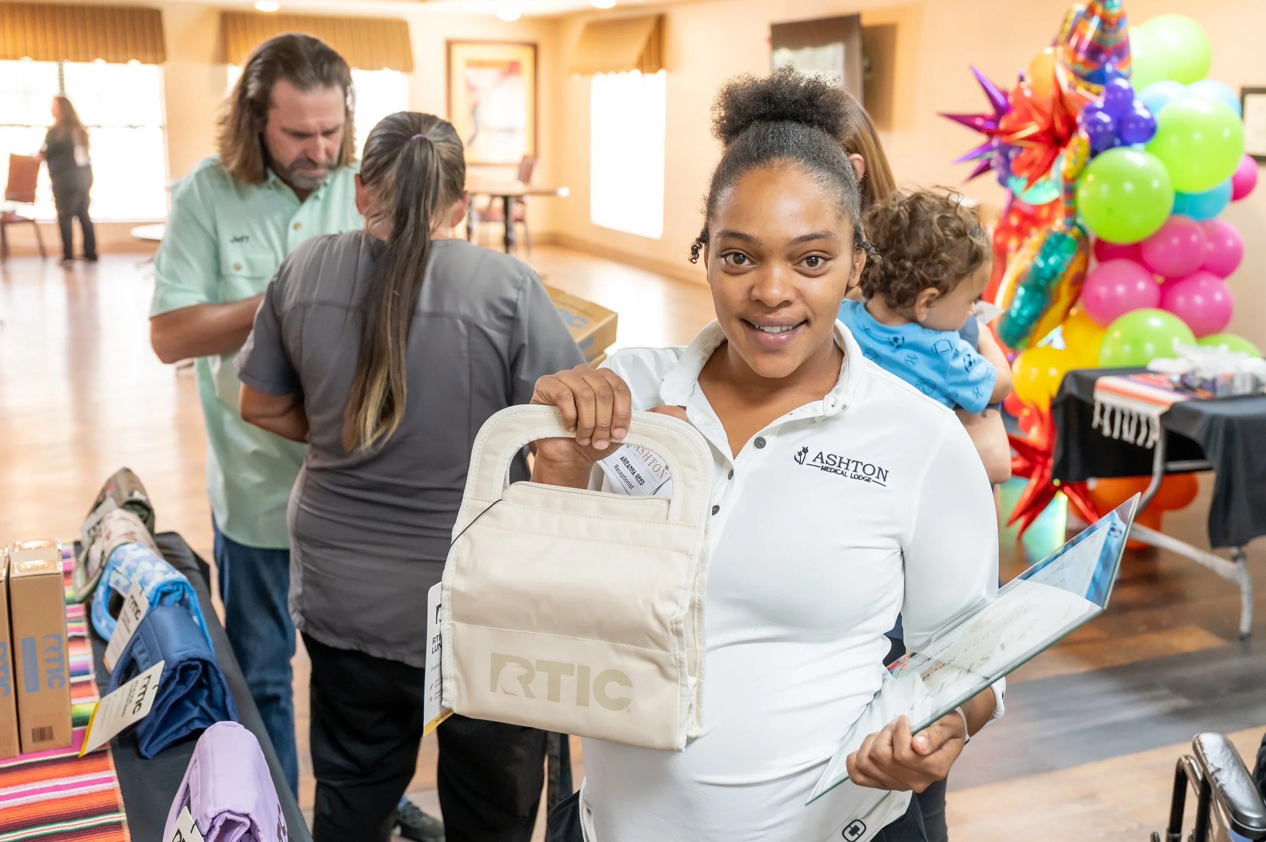 A woman with a dark skin tone and curly hair, wearing a white Ashton Medical Lodge shirt, is smiling and holding a beige tote bag and a clear folder, standing at a community event with colorful balloons and tables in the background.