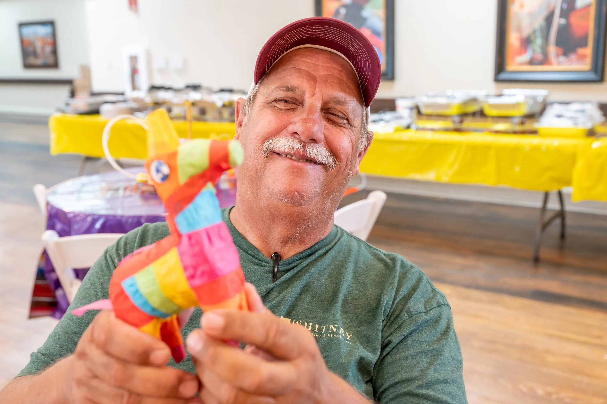 Smiling man in a green shirt and red cap holding a colorful, patchwork llama plush toy at a festive gathering with yellow and purple tablecloths in the background.