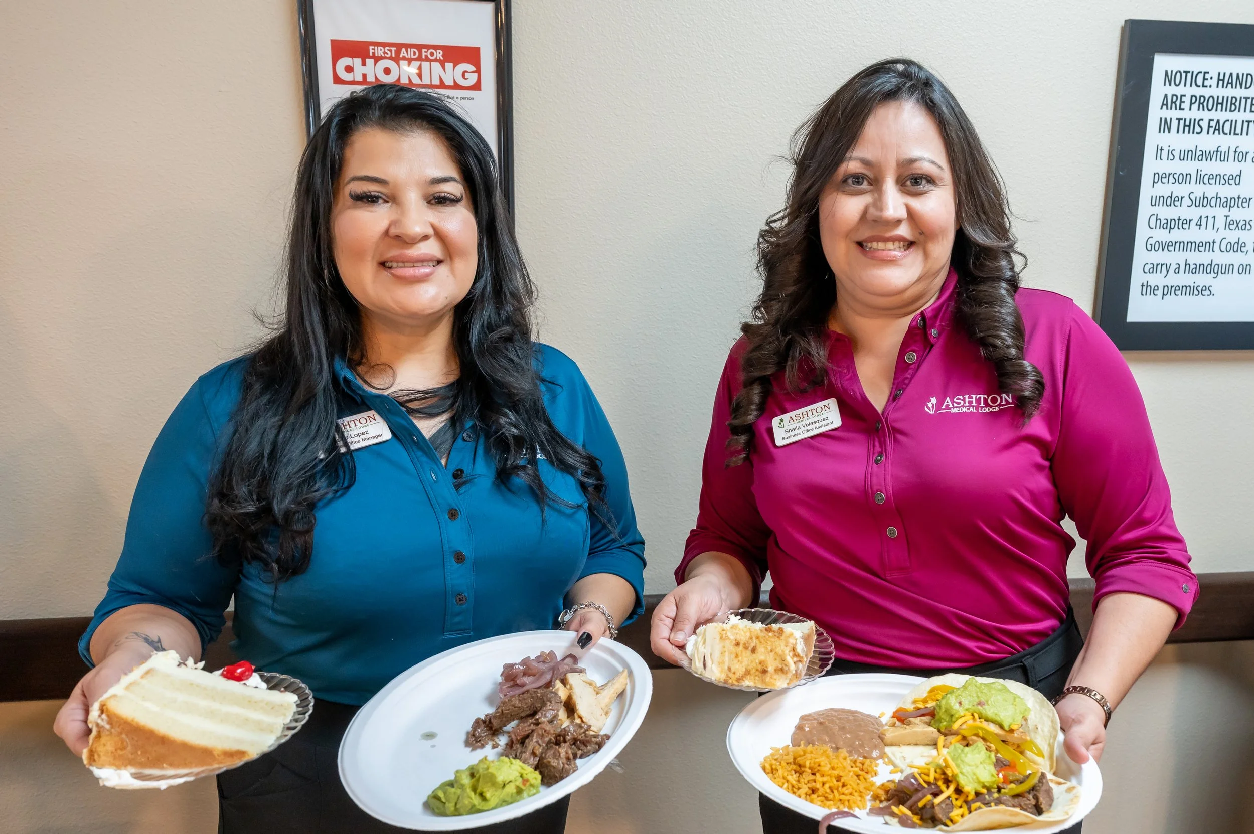 Two women wearing purple and blue shirts with name tags holding plates of food, standing in a room with framed notices on the wall.