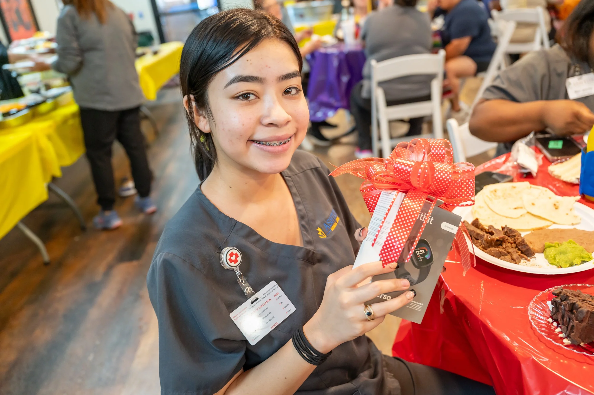 A young woman in a gray uniform smiling at the camera, holding a gift box with a red bow, sitting at a table with plates of food, at a celebration or event.