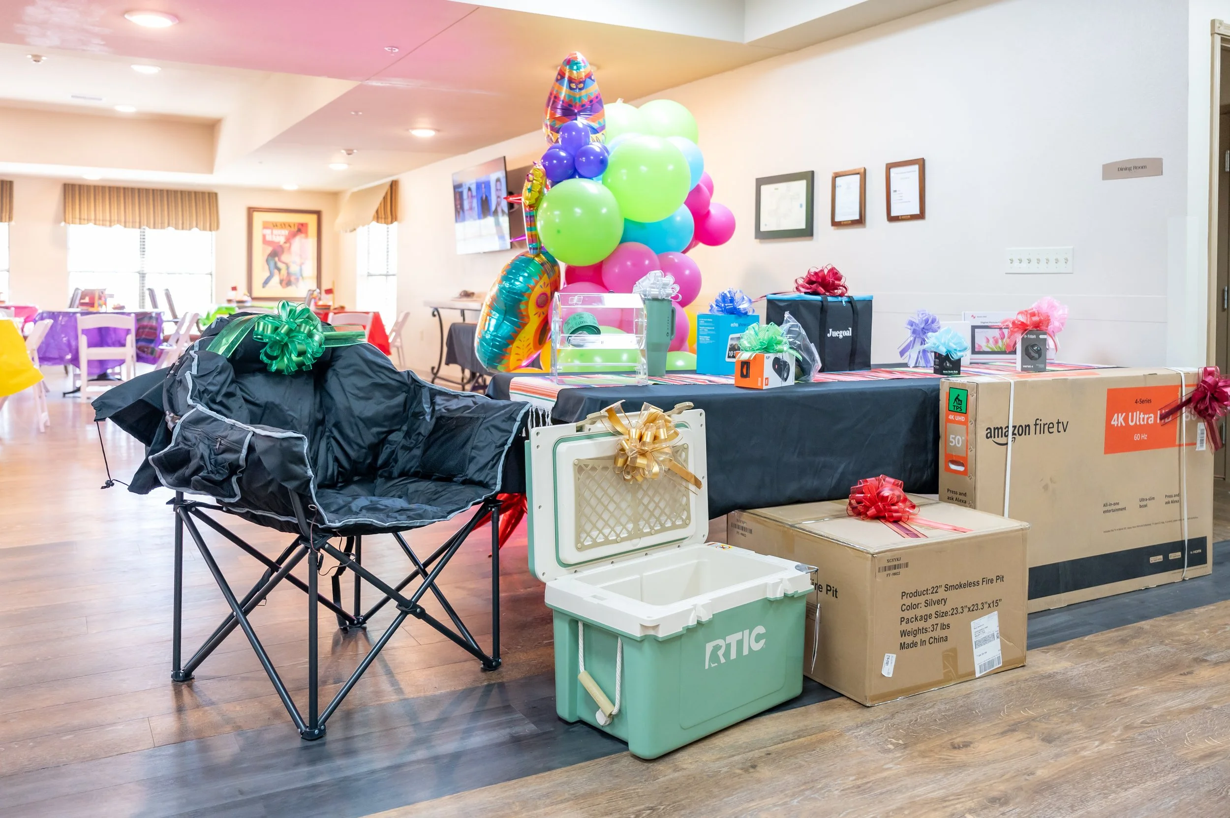 Party room with tables, chairs, colorful balloons, gifts, and a cooler, likely set up for a celebration.