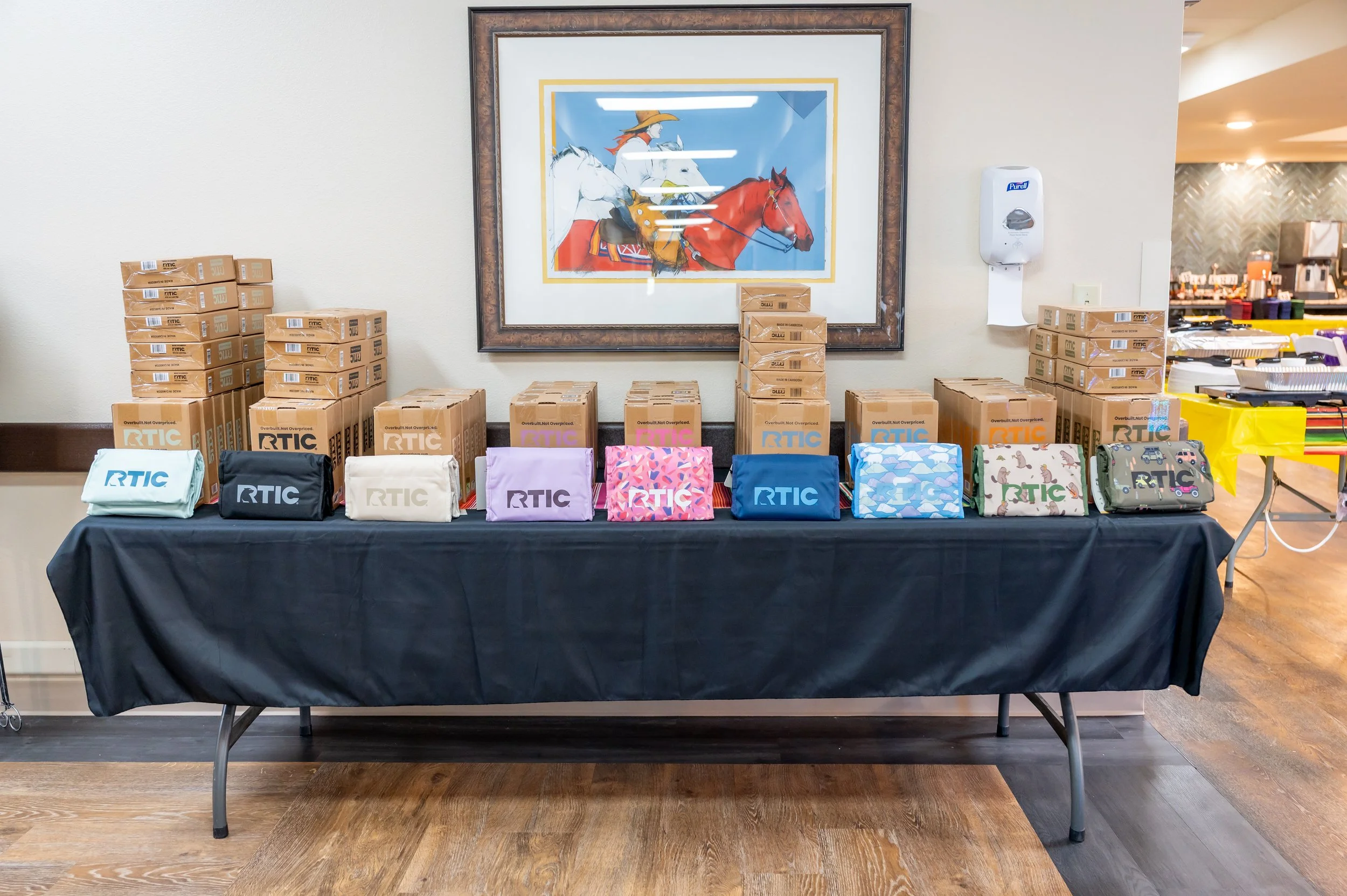 Table covered with a black cloth displaying various colorful bags with the RTIC logo, along with stacked boxes in the background. A painting of a woman riding a white horse is hanging on the wall above the table. To the right, a wall-mounted soap dis