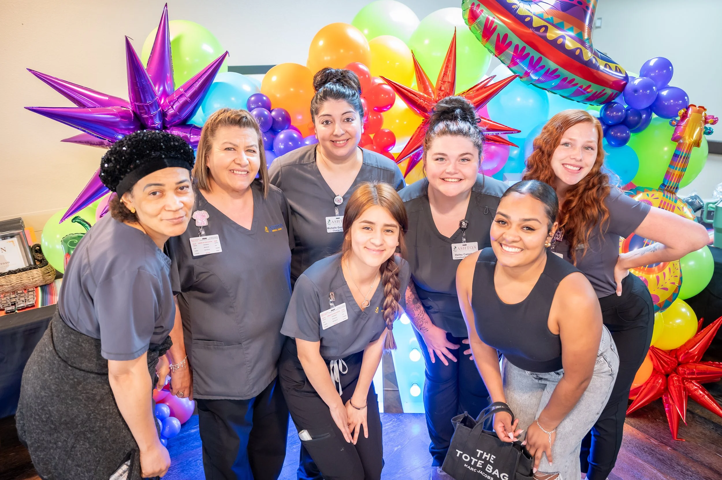 Group of seven women, some in grey scrubs, gathered together smiling in front of colorful balloons and decorations, celebrating a festive occasion.