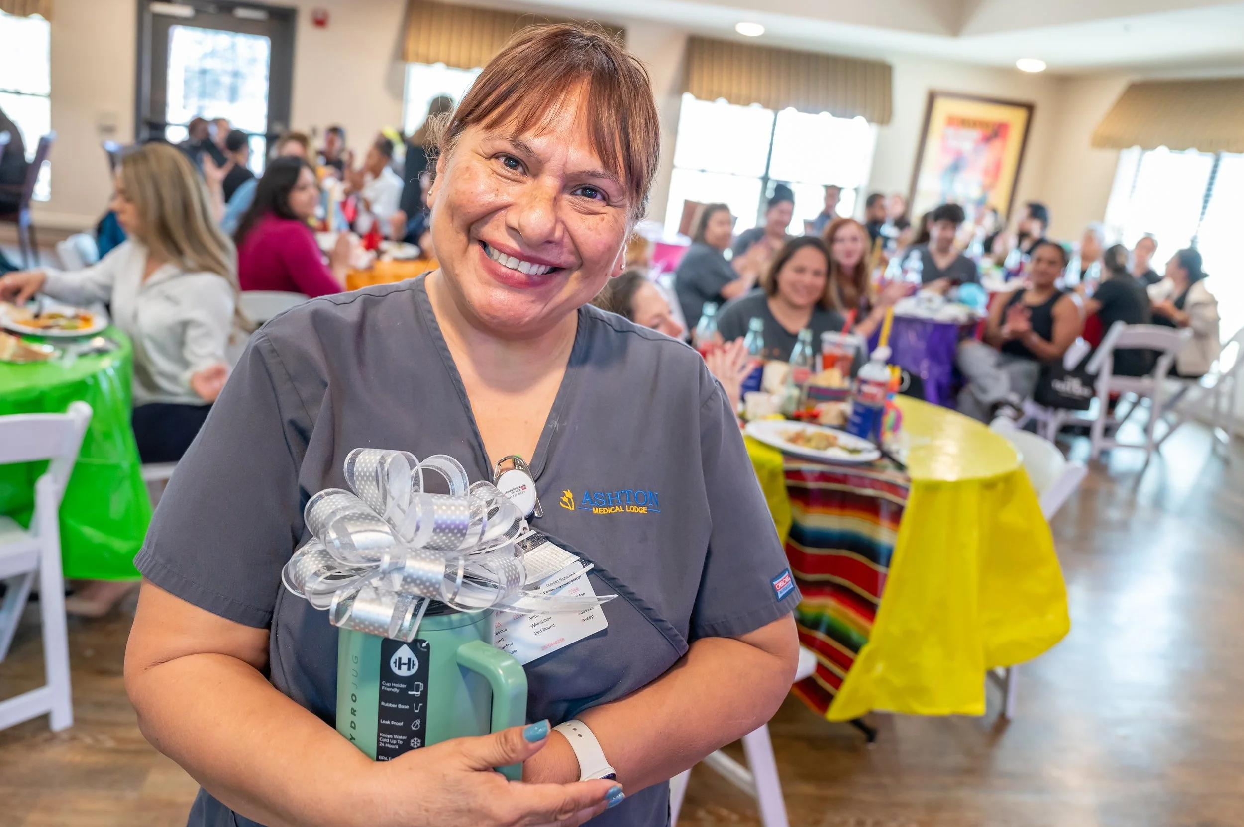 A woman in gray medical scrubs holding a gift with a bow while smiling, in a brightly decorated room filled with seated people at tables.