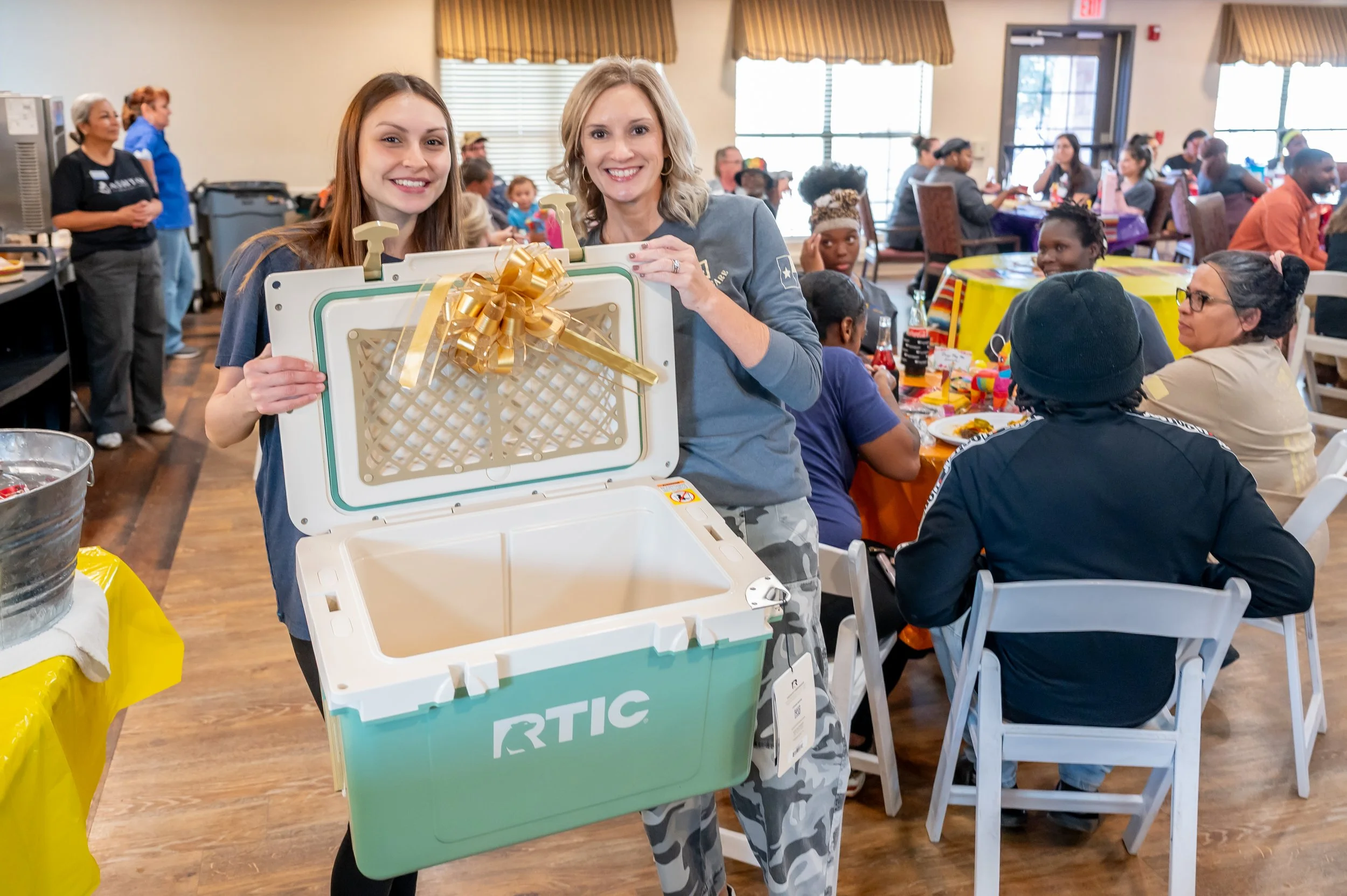 Two women are smiling and holding an open cooler with a gold bow inside at a crowded indoor celebration or gathering.