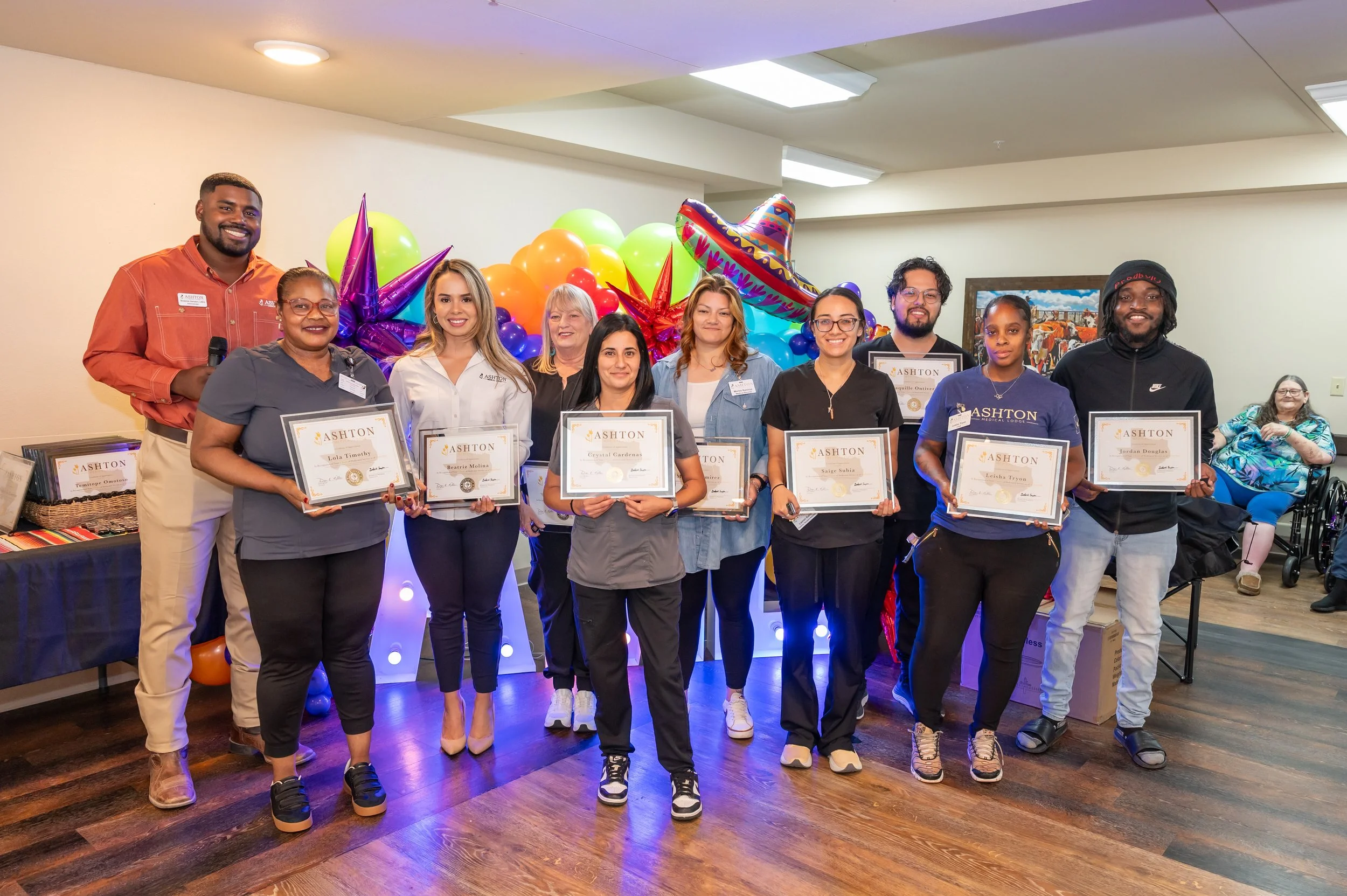 A group of ten people standing indoors, holding certificates, with colorful balloons and decorations behind them, celebrating an award or achievement.