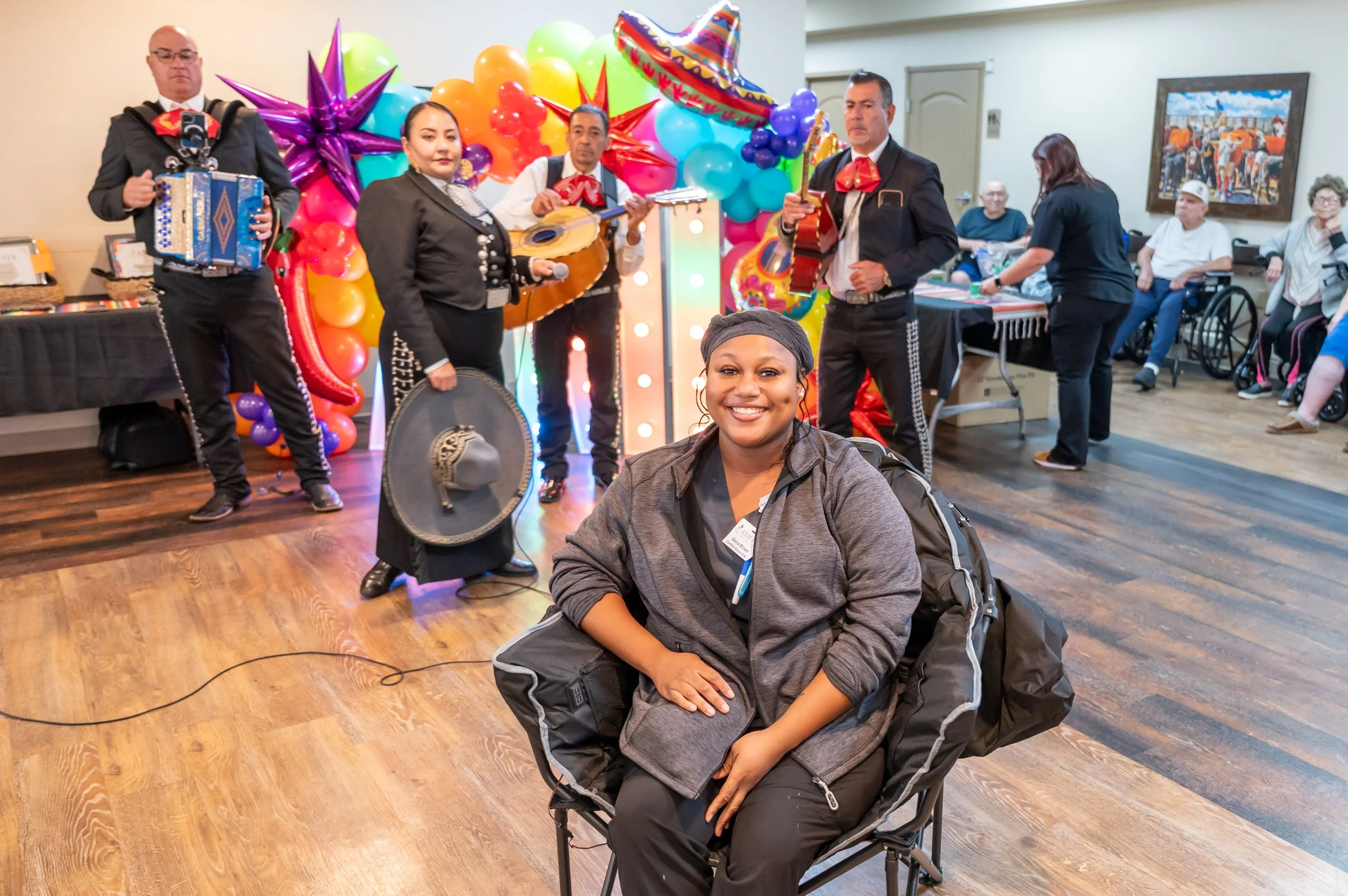 A group of musicians performing at a festive celebration with colorful balloons and decorations in the background, and people seated in the audience, including a woman smiling in the foreground.