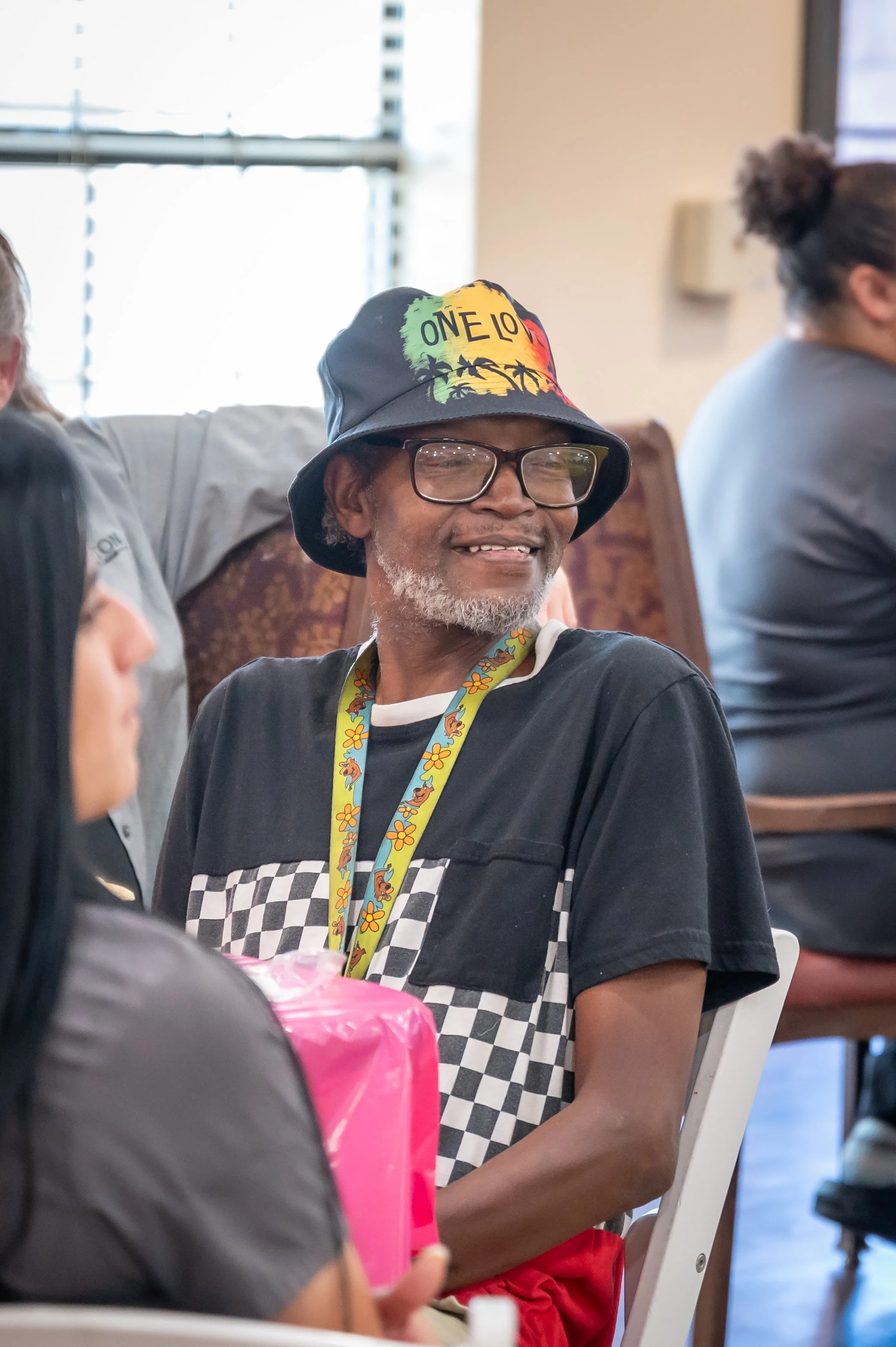 Older man with a beard and glasses, wearing a colorful bucket hat, sitting at a gathering, smiling, with other people around.