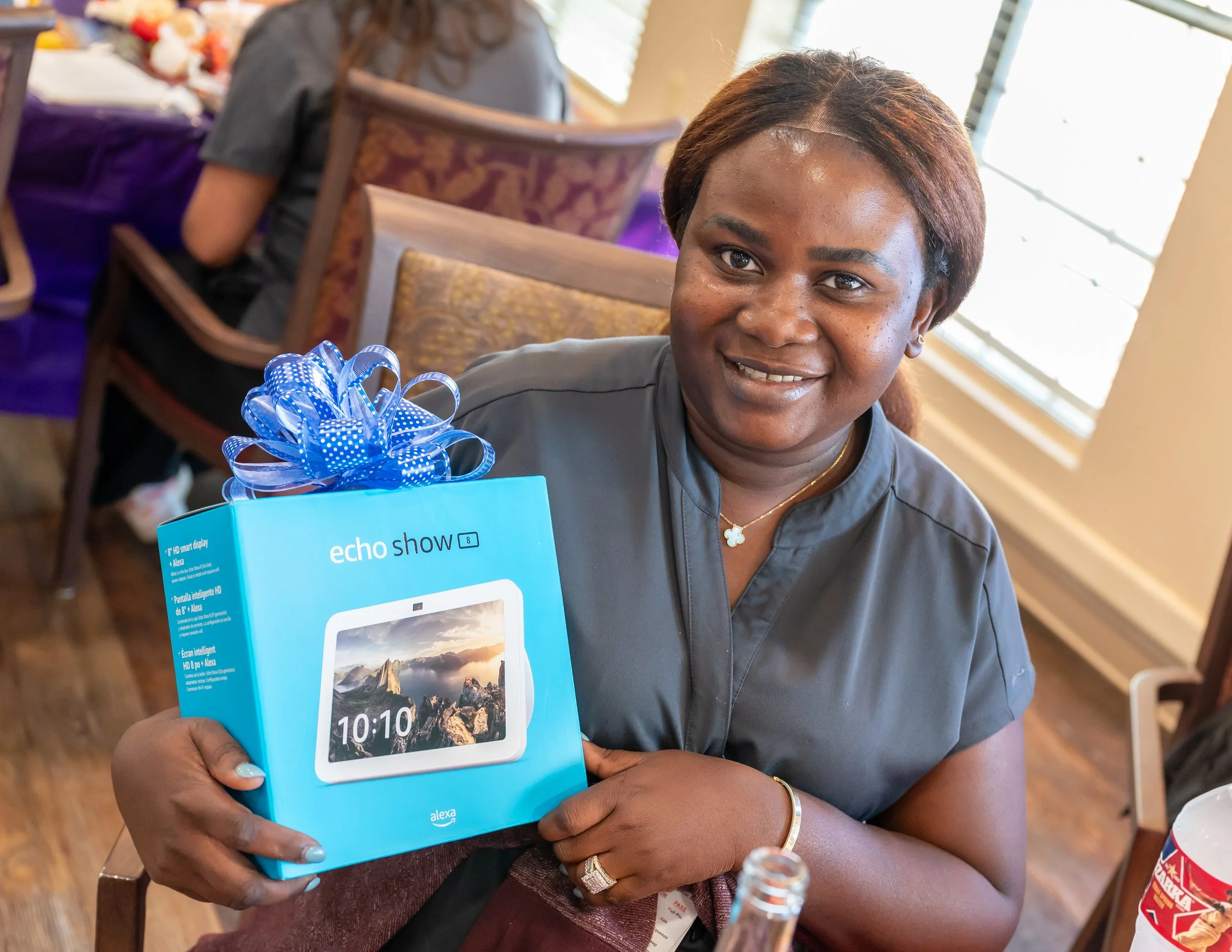 A woman in a gray shirt holding a blue box with a smart display, smiling at the camera, in a room with other people and a window in the background.