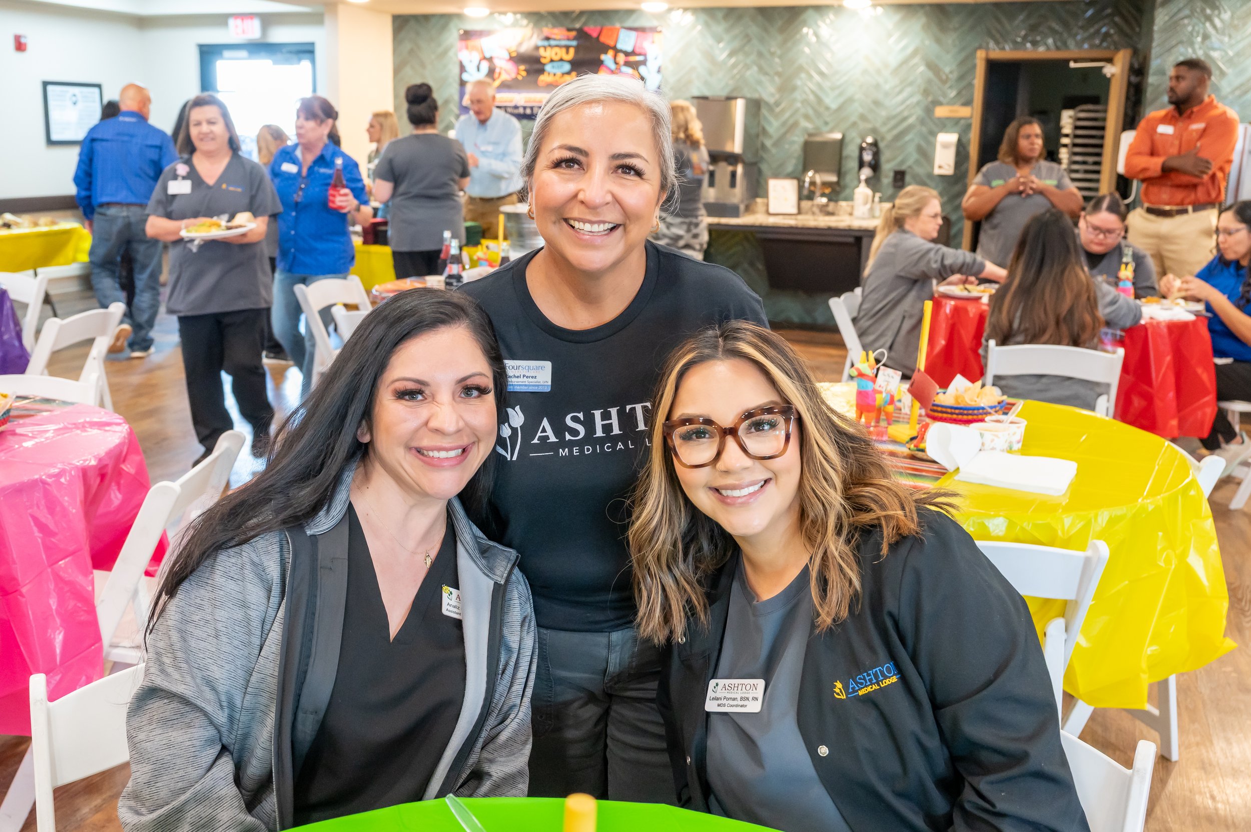 Three women smiling at a social gathering. One woman in the middle standing behind two women seated at a table, all three wearing ASHTON Medical Group shirts or jackets. The event has colorful tables with decorations, and people are mingling and eati
