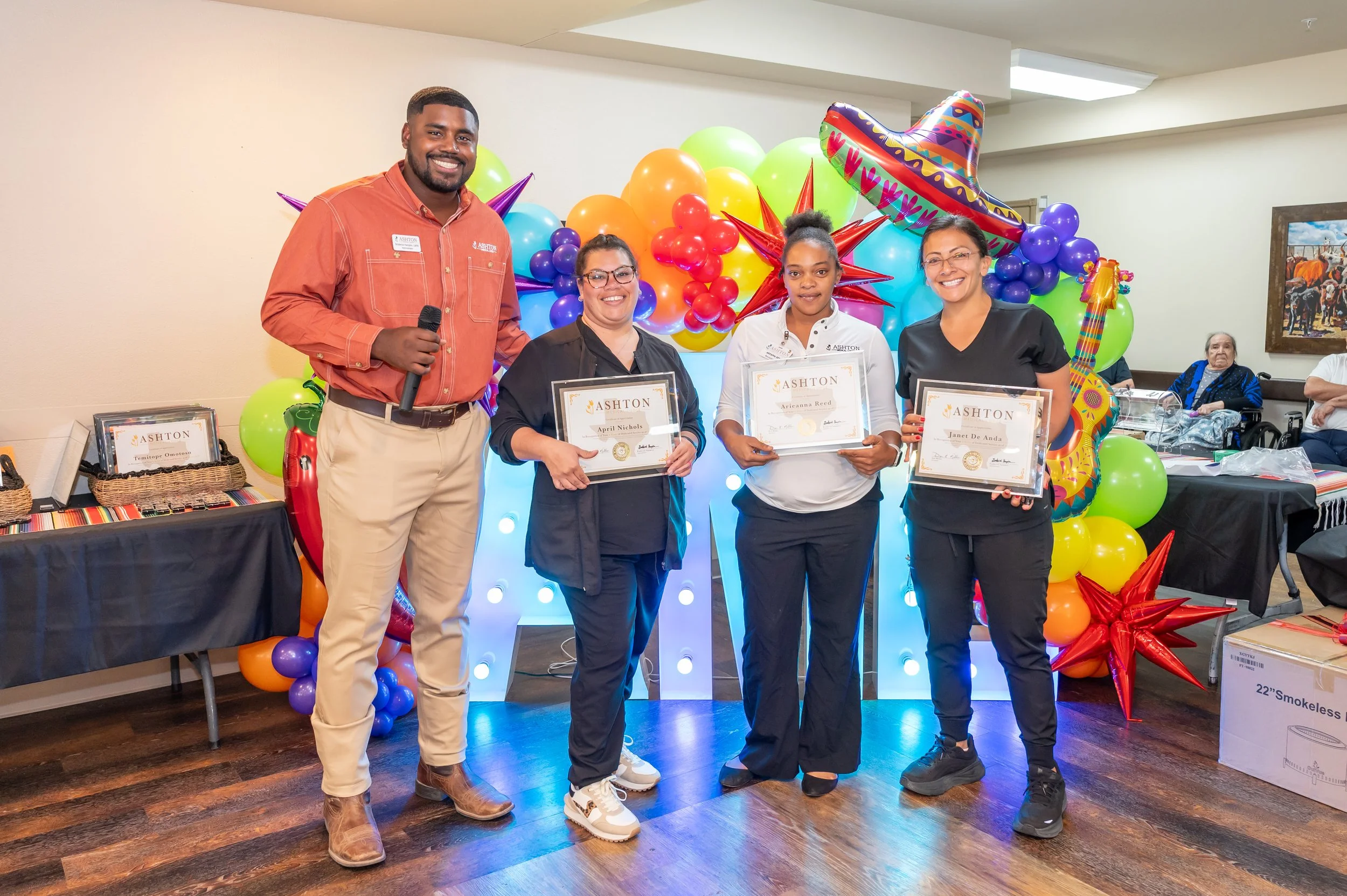 Four people standing in front of colorful balloons and decorations, holding certificates, at an award ceremony or celebration event.