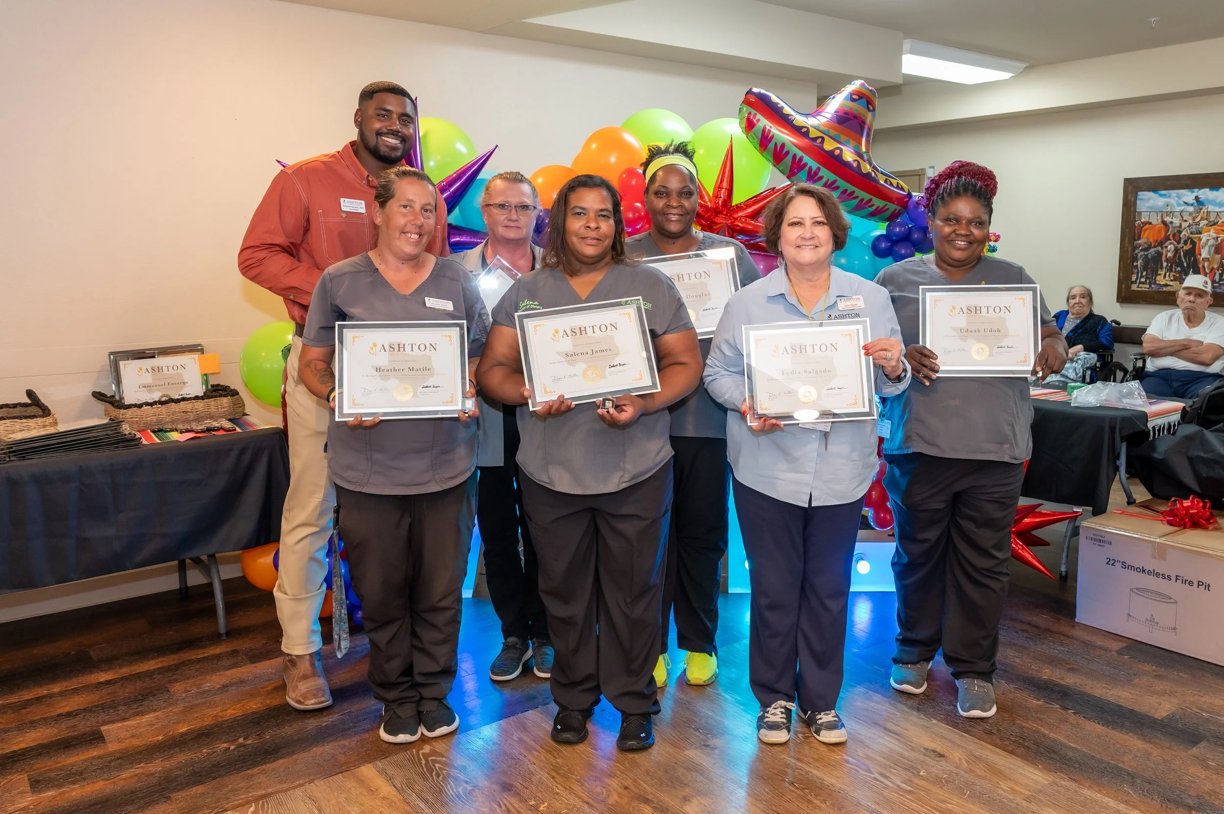 Group of six women and one man posing for a photo, holding certificates, at an award ceremony with colorful balloons and decorations in the background.
