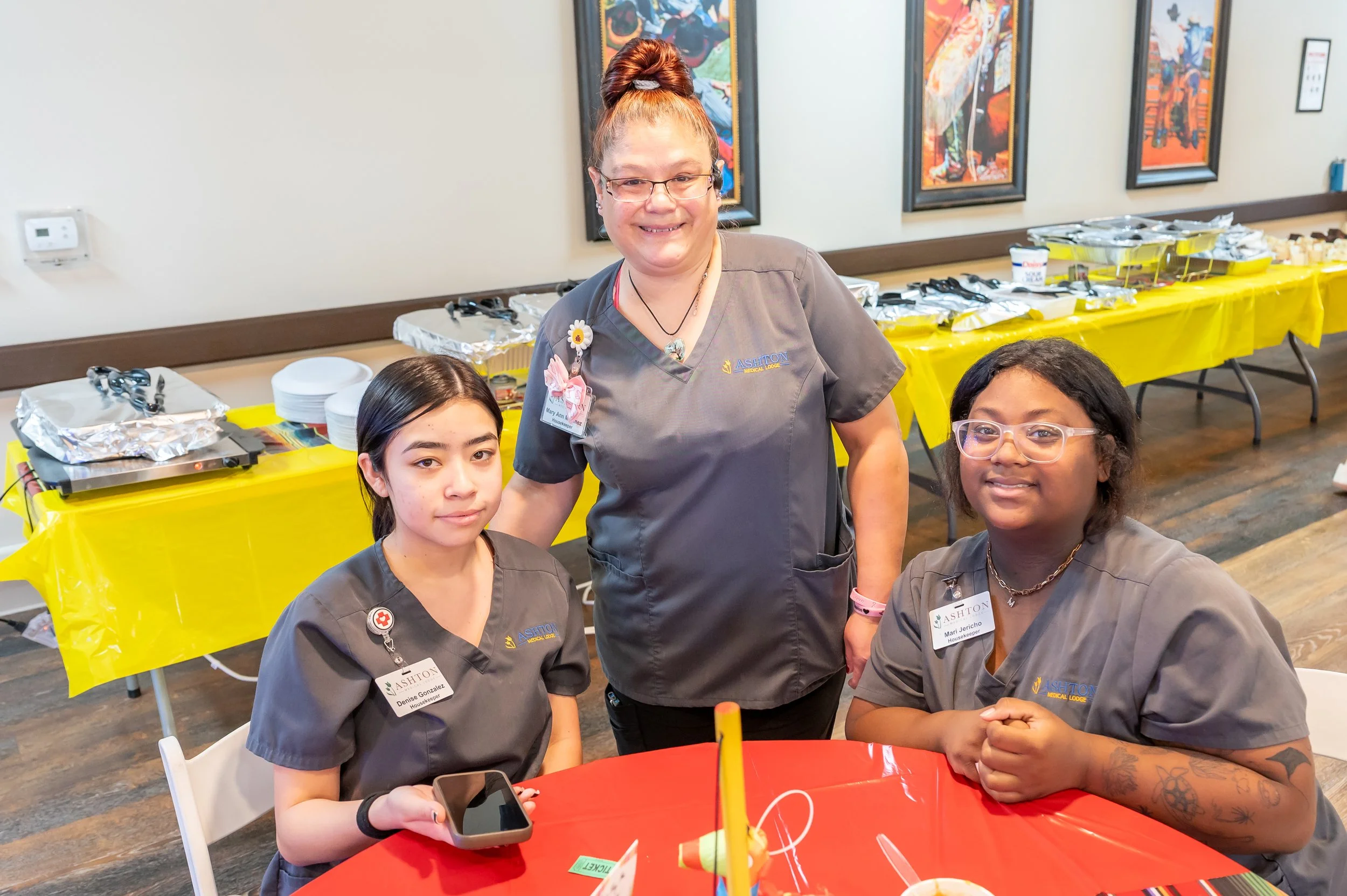 Three women in gray scrubs smiling at a gathering. Two women are seated at a red table, and one woman is standing behind them. Background shows a long table with food in foil and dishes, and colorful artwork on the wall.