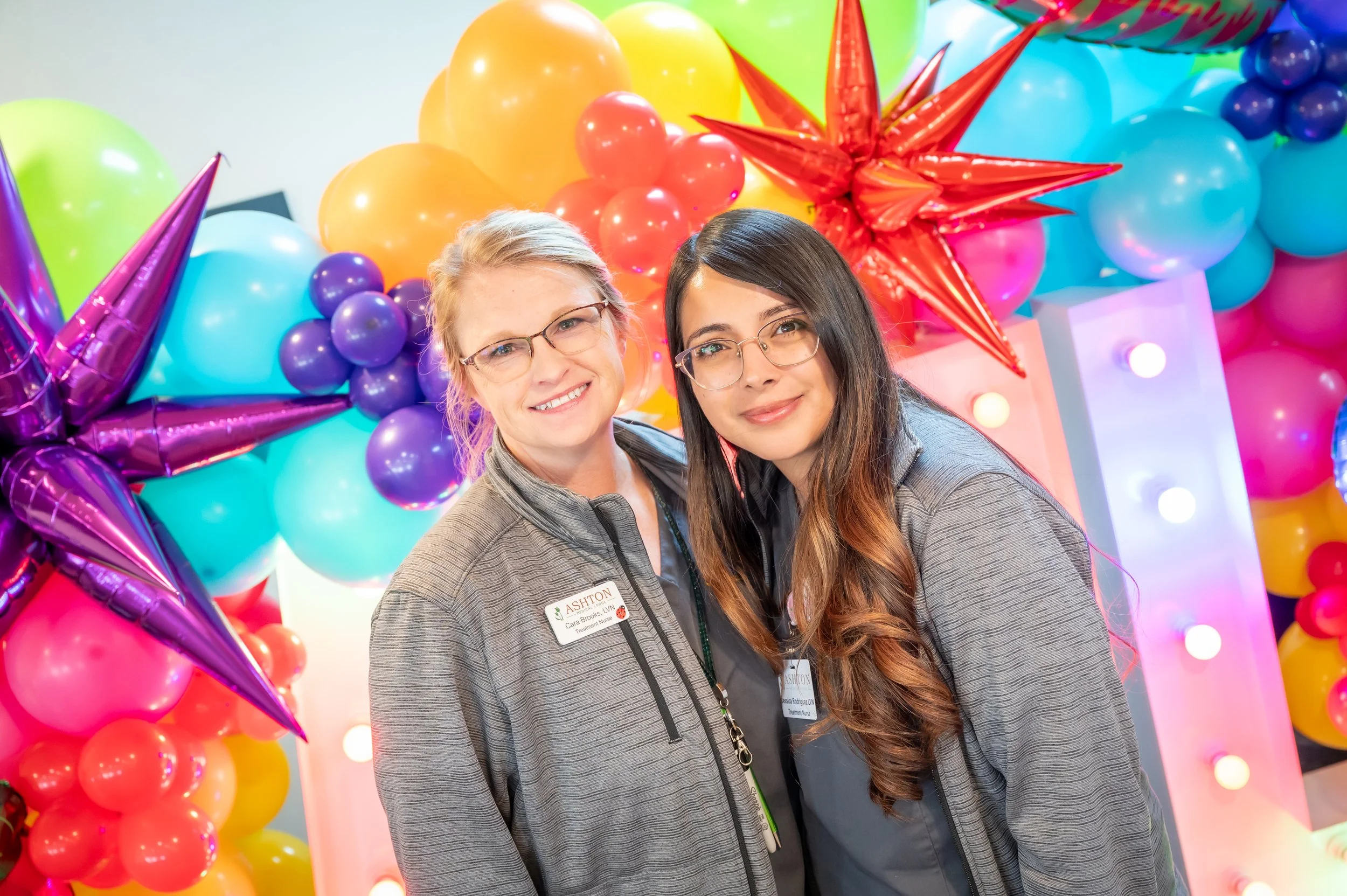 Two women smiling in front of colorful balloon decorations with large stars and lights.