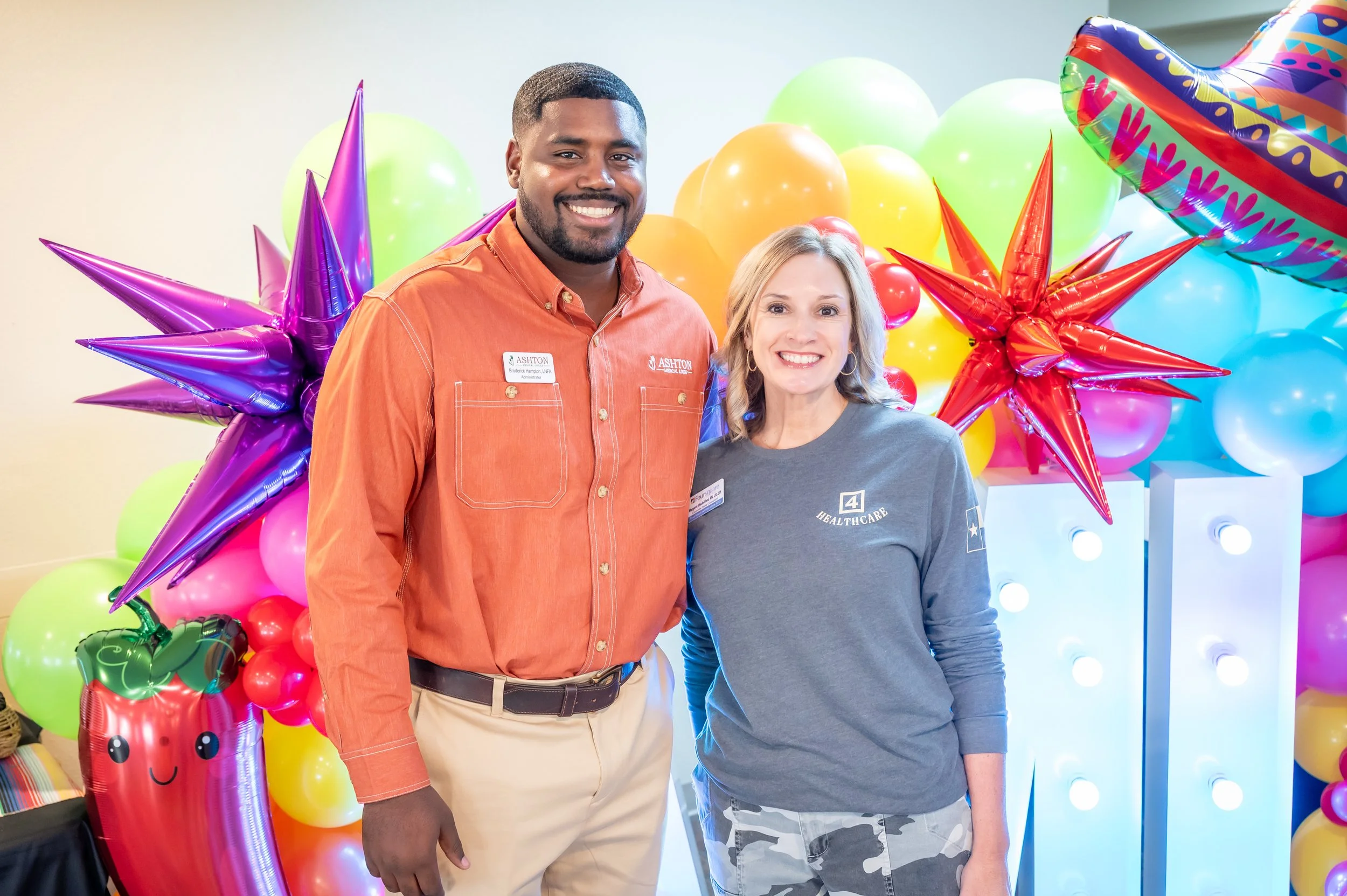 Two smiling people standing together in front of colorful balloons and decorations at a celebration or event.