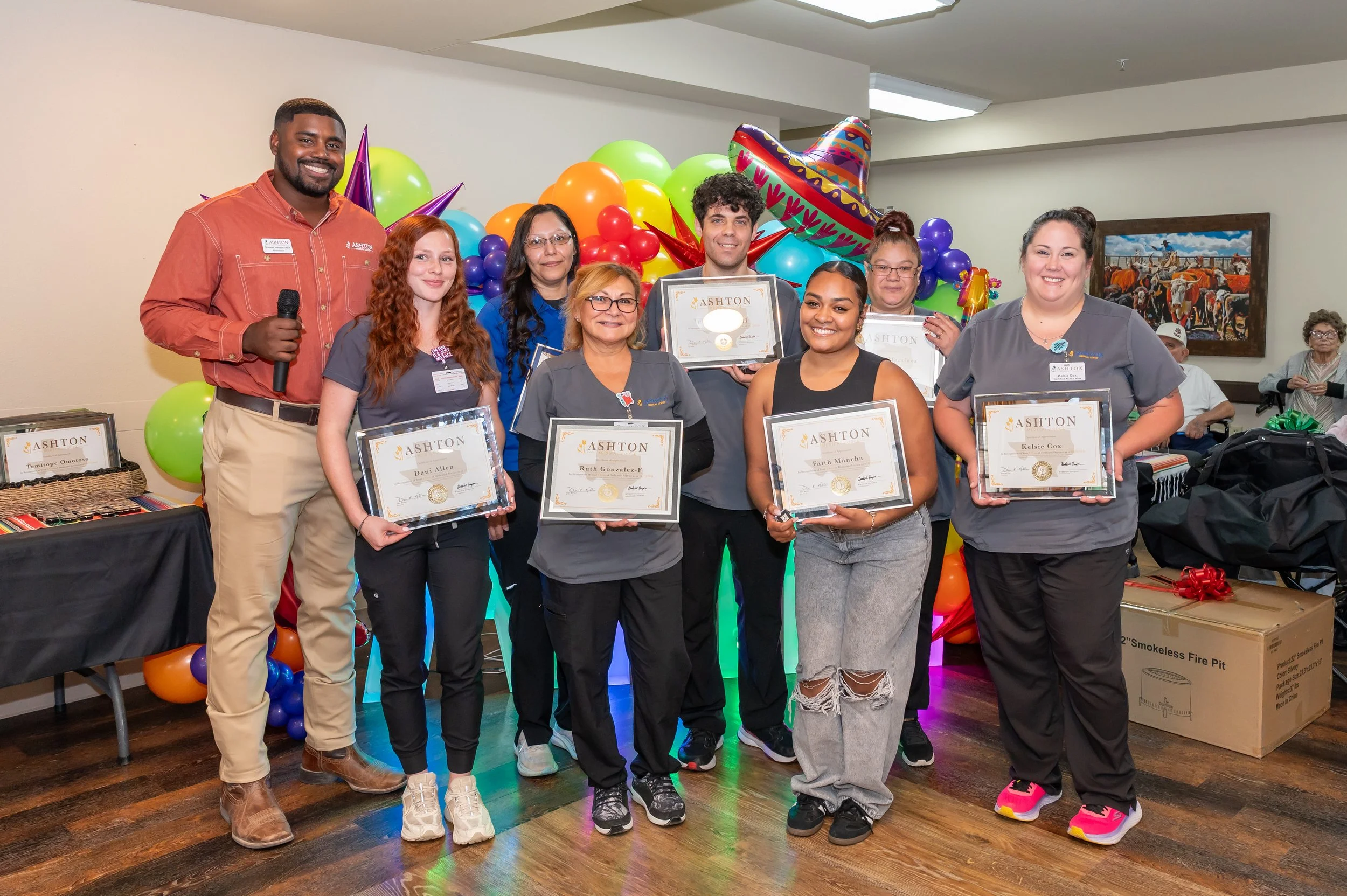 Group of eight people standing indoors, holding framed certificates, with colorful balloons and a sombrero decoration in the background, celebrating an achievement or event.