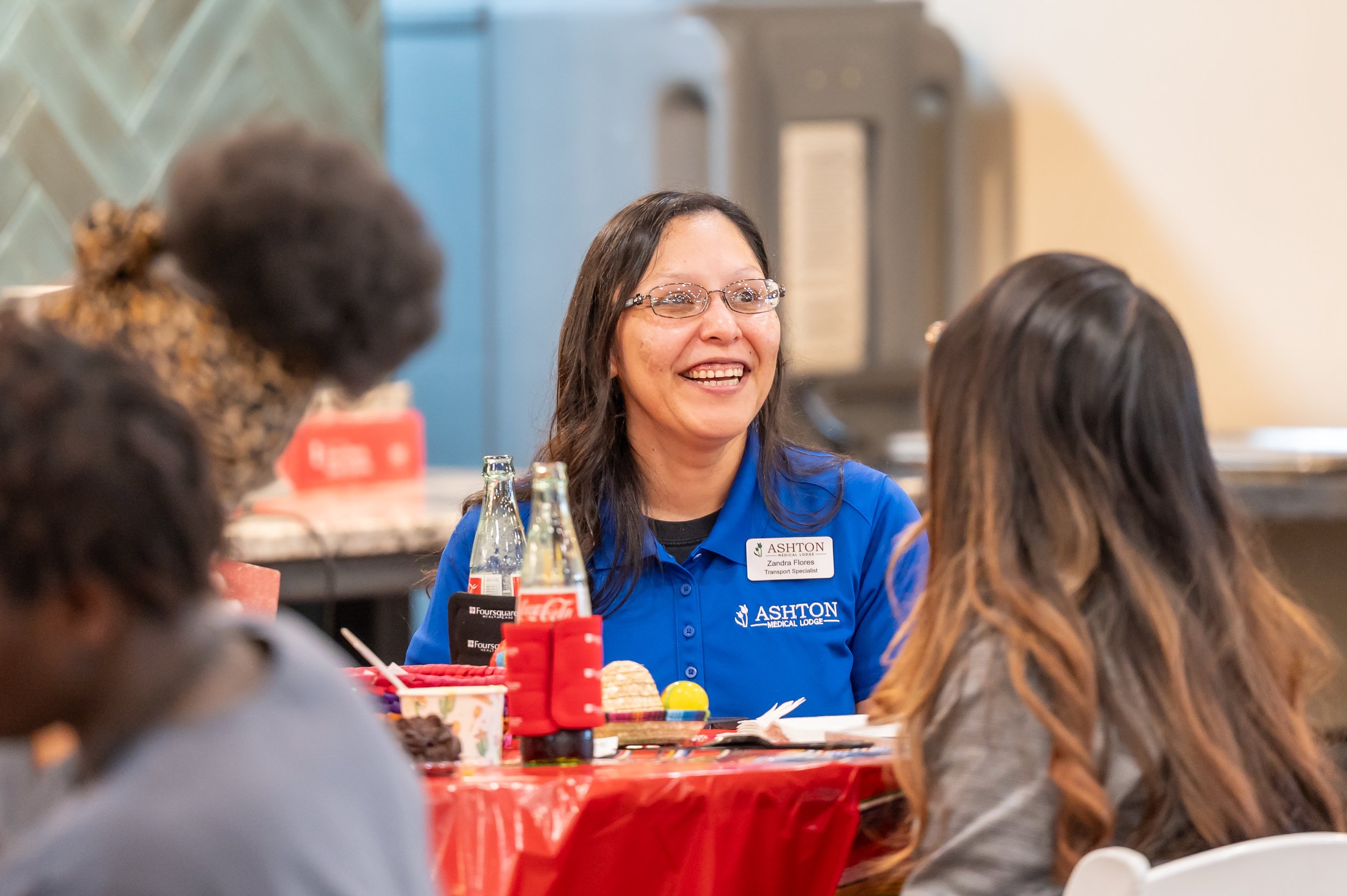A woman with glasses, wearing a blue shirt with a name tag that reads 'Zandra Flores', is smiling and talking to two young women at a holiday gathering. There are bottles, food, and decorations on the table.