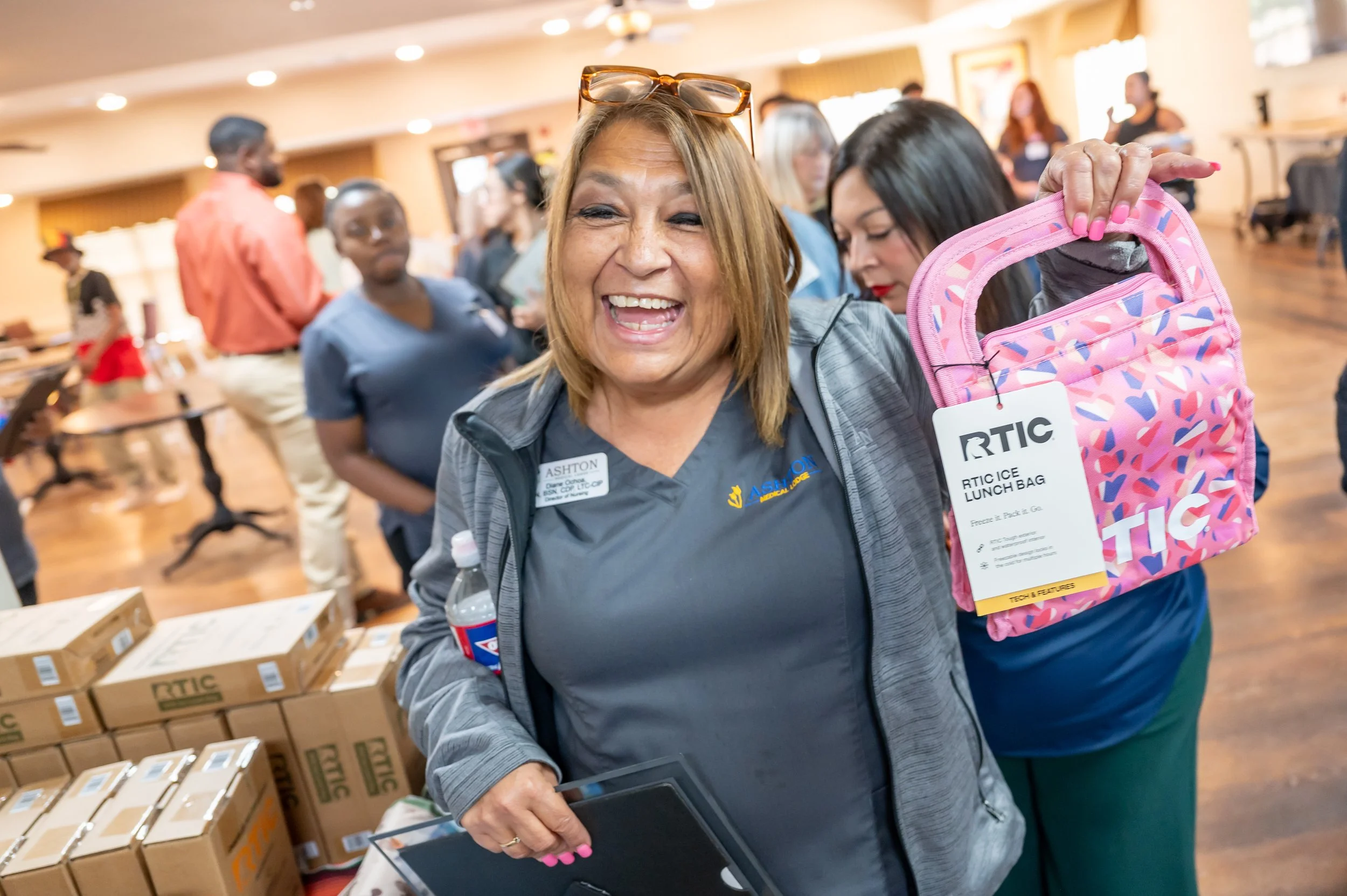 A smiling woman holding a pink RTIC lunch bag in a crowded indoor setting, surrounded by boxes and other people.