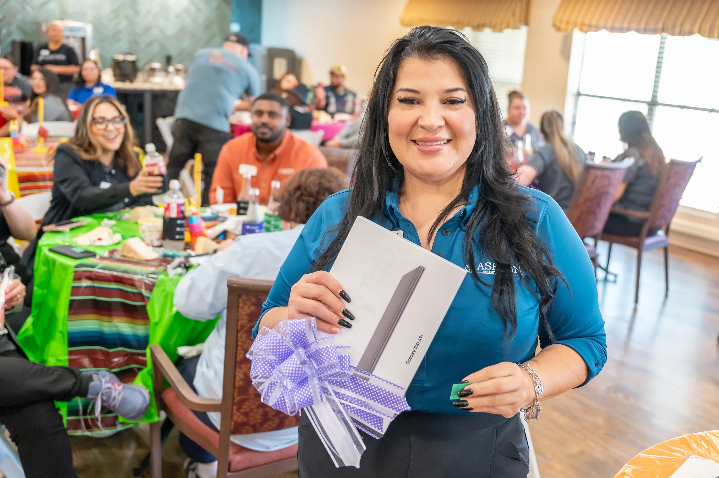 Woman holding a boxed electronic device at a social gathering or celebration, with people sitting at tables in the background.