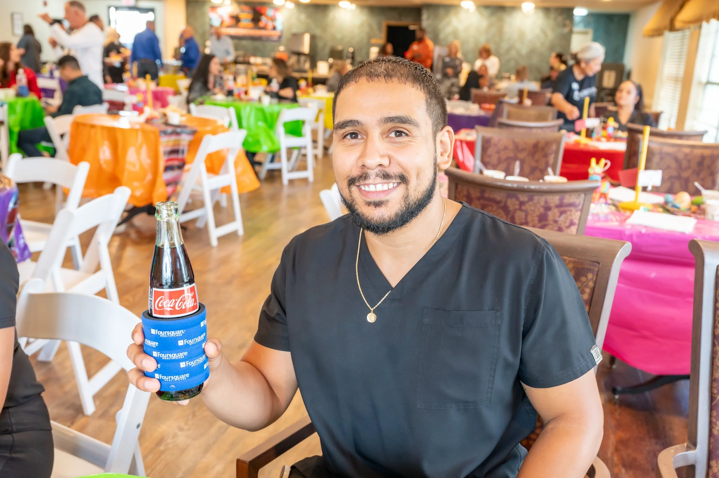 A man smiling and holding a bottle of Coca-Cola in a colorful party setting with decorated tables and people in the background.