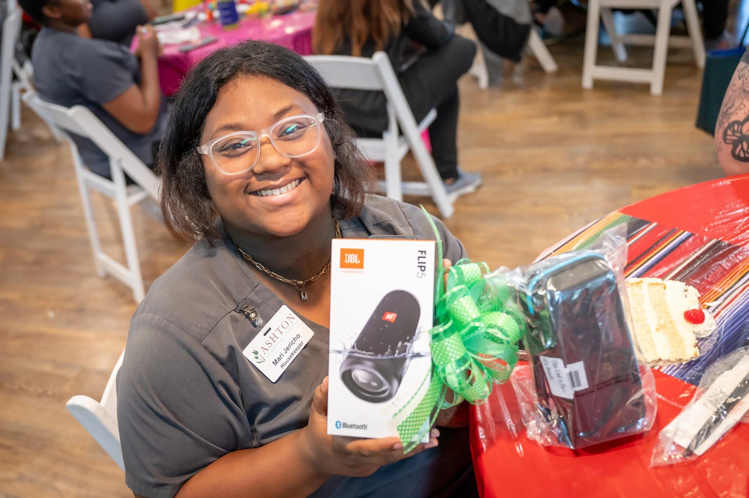 A smiling woman with glasses and a name tag holding a JBL Flip 5 waterproof portable Bluetooth speaker, sitting at a decorated table with cake slices, at a celebration event.