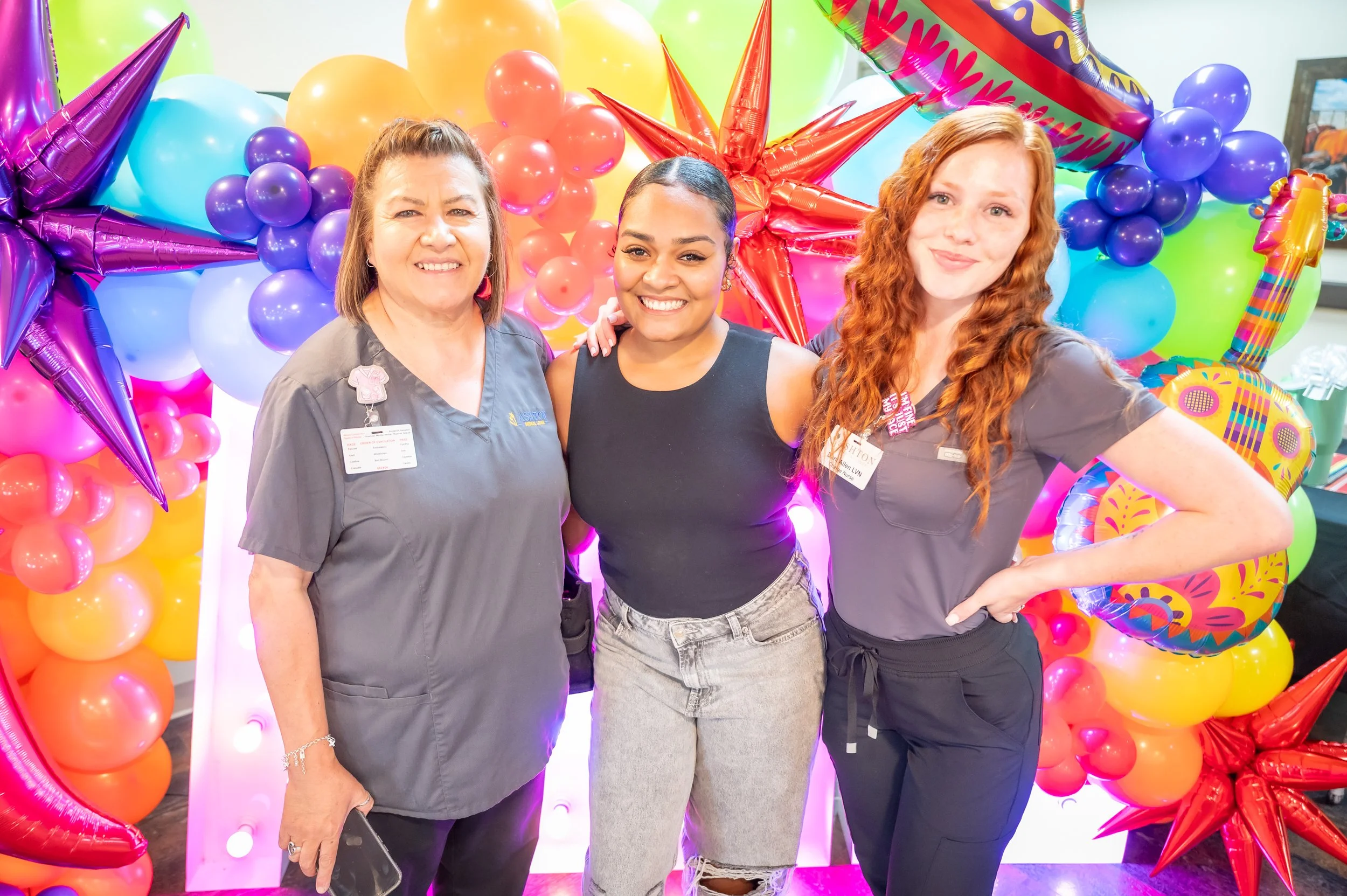 Three women standing together in front of colorful balloons and star-shaped decorations at a celebration or party.