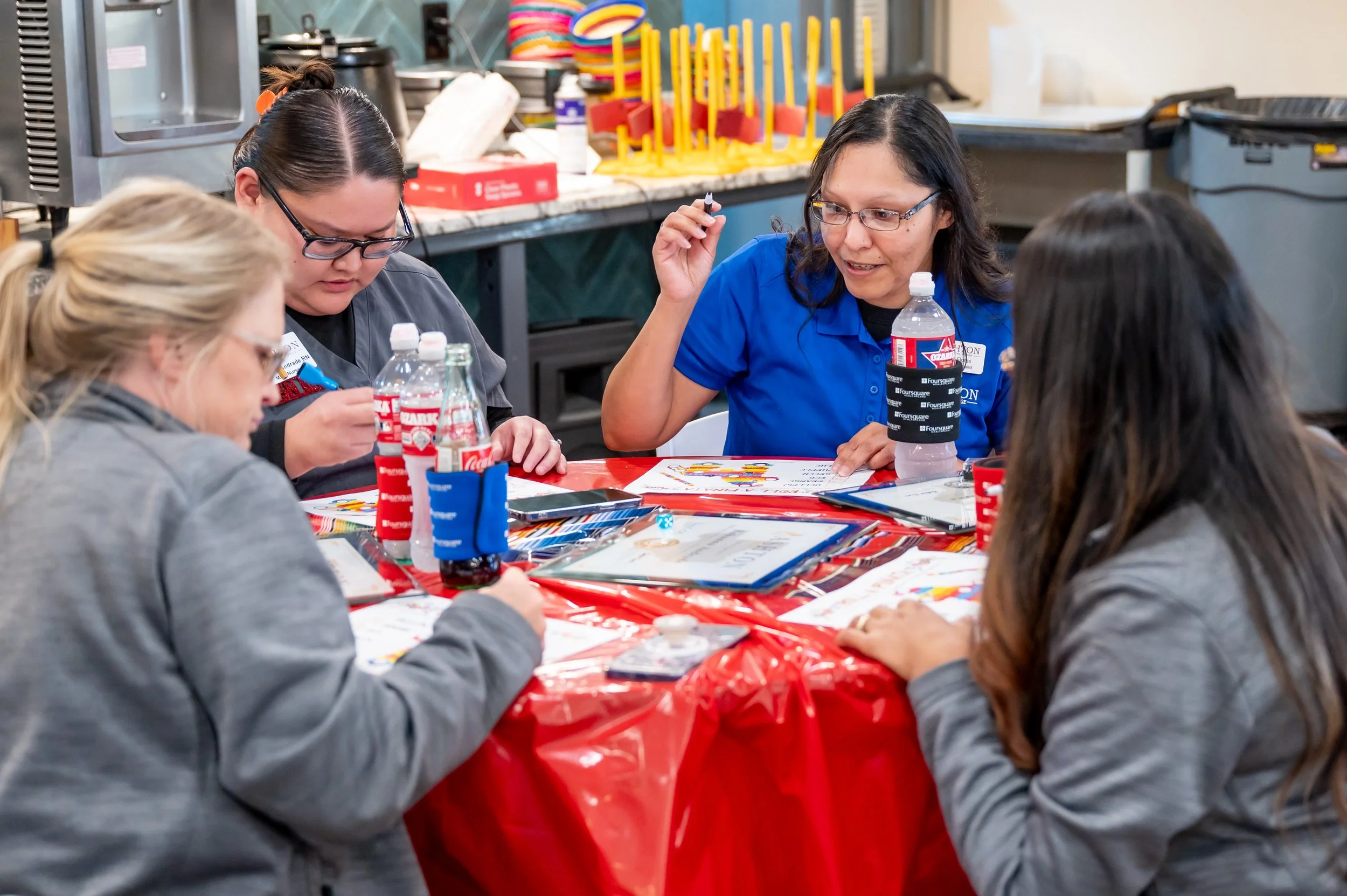 Four women sitting around a table with red tablecloth, engaged in a discussion or activity. They are in a kitchen or cafeteria setting, with bottles of water, paperwork, and small items on the table.