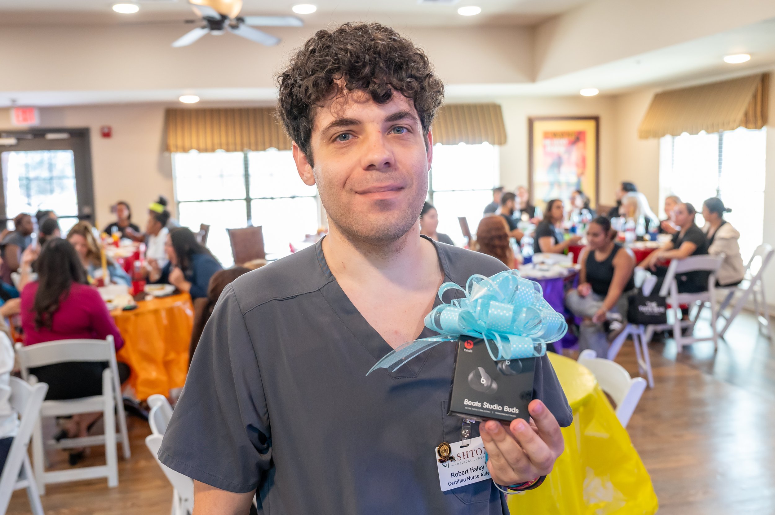 A young man with curly brown hair and a gray uniform holding a blue ribboned gift box at a buffet-style gathering with many seated people in the background.