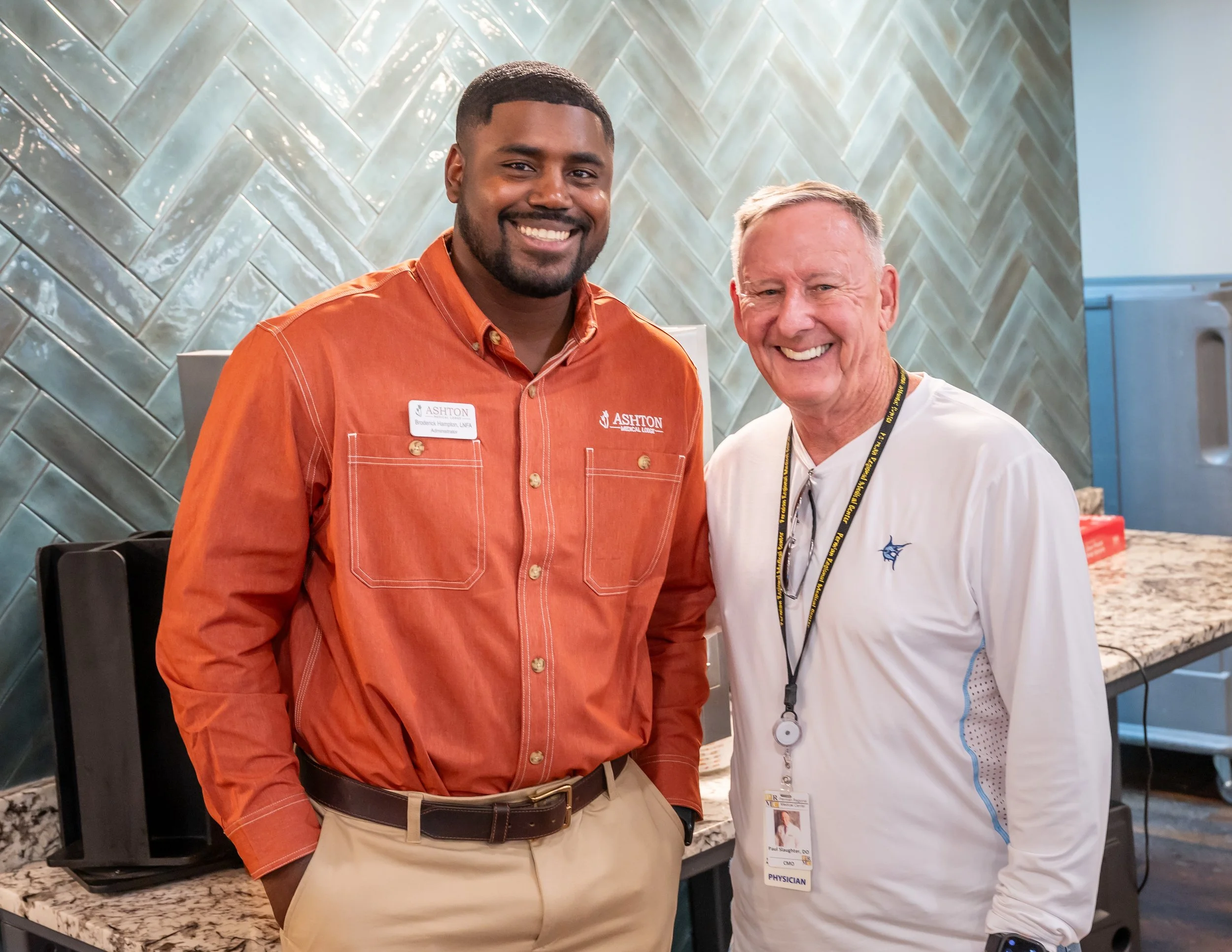 Two men smiling and posing together indoors, one wearing an orange shirt with a name badge and the other wearing a white shirt with a lanyard and badge, standing in front of a tiled wall and marble countertop.
