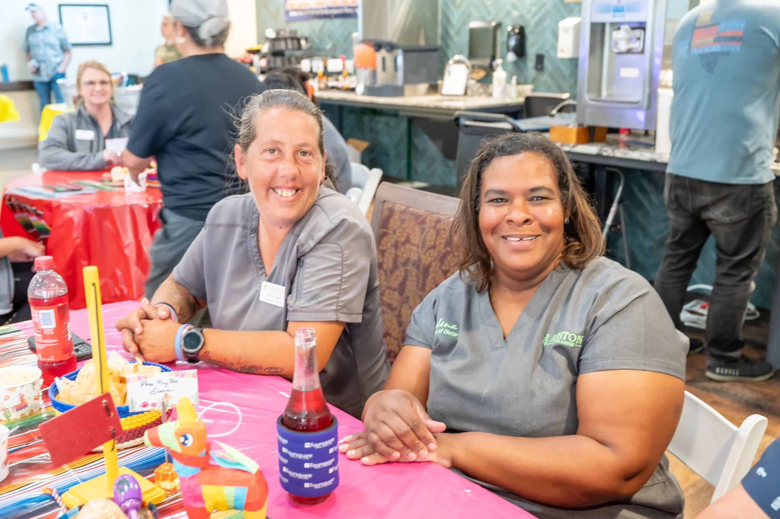 Two women wearing gray scrubs, sitting at a table with colorful decorations and food, smiling at the camera at a celebration event.