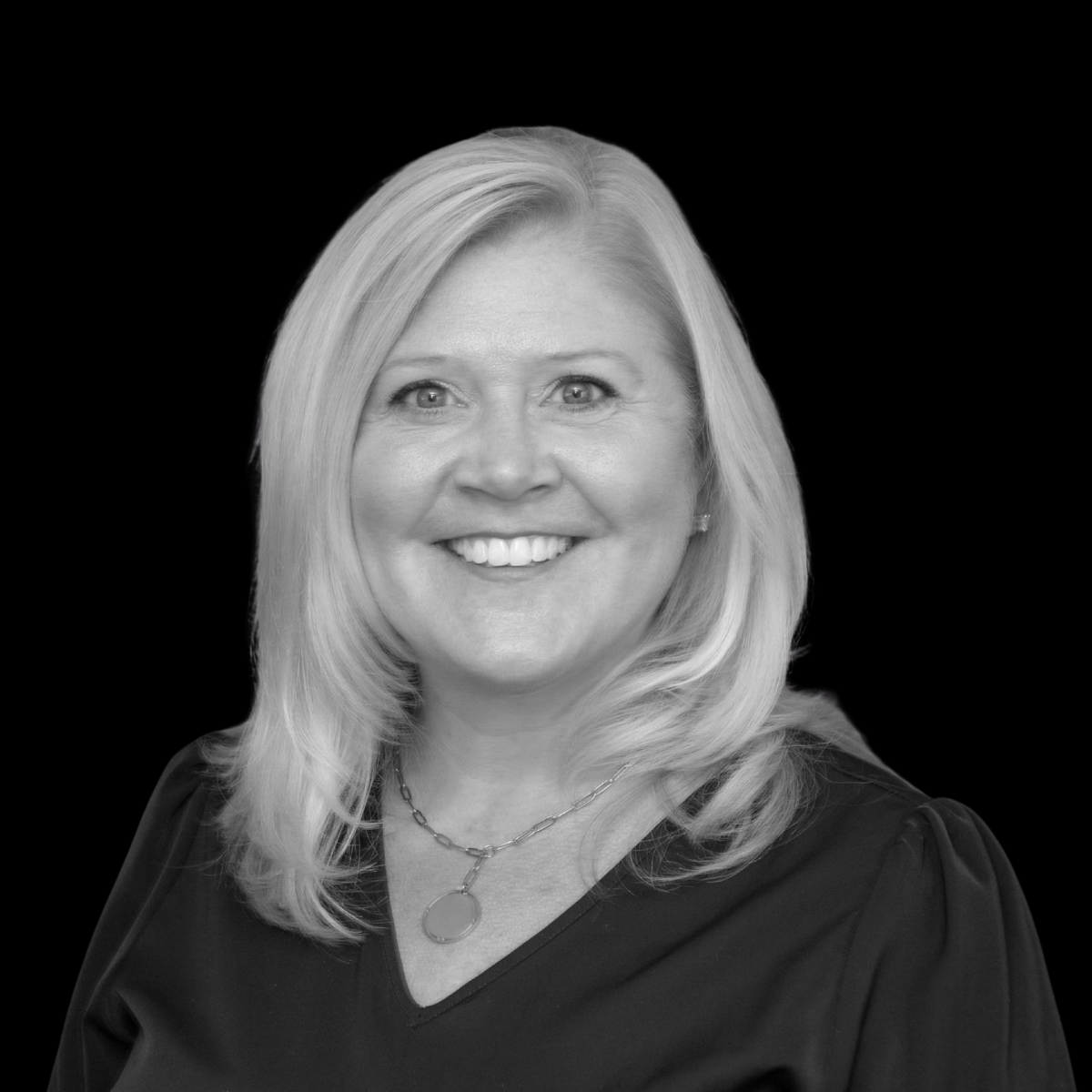 Black and white portrait of a smiling woman with shoulder-length blonde hair, wearing a black top and a necklace with a circular pendant, against a dark background.