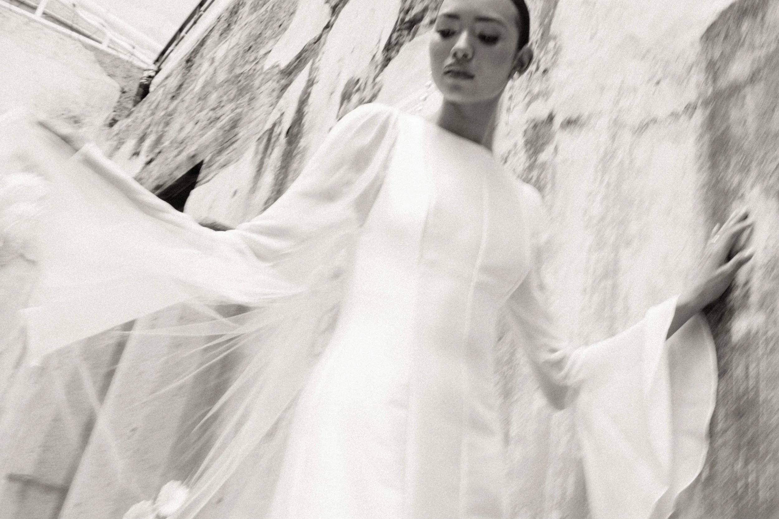A woman in a flowing white dress posed against a textured stone wall in black and white.