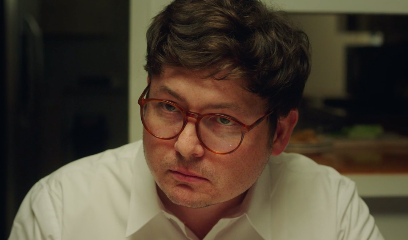 Still from the short film, EXTERMINATOR. A man (Allan McLeod) wearing glasses and a white shirt stares at his dinner guests.