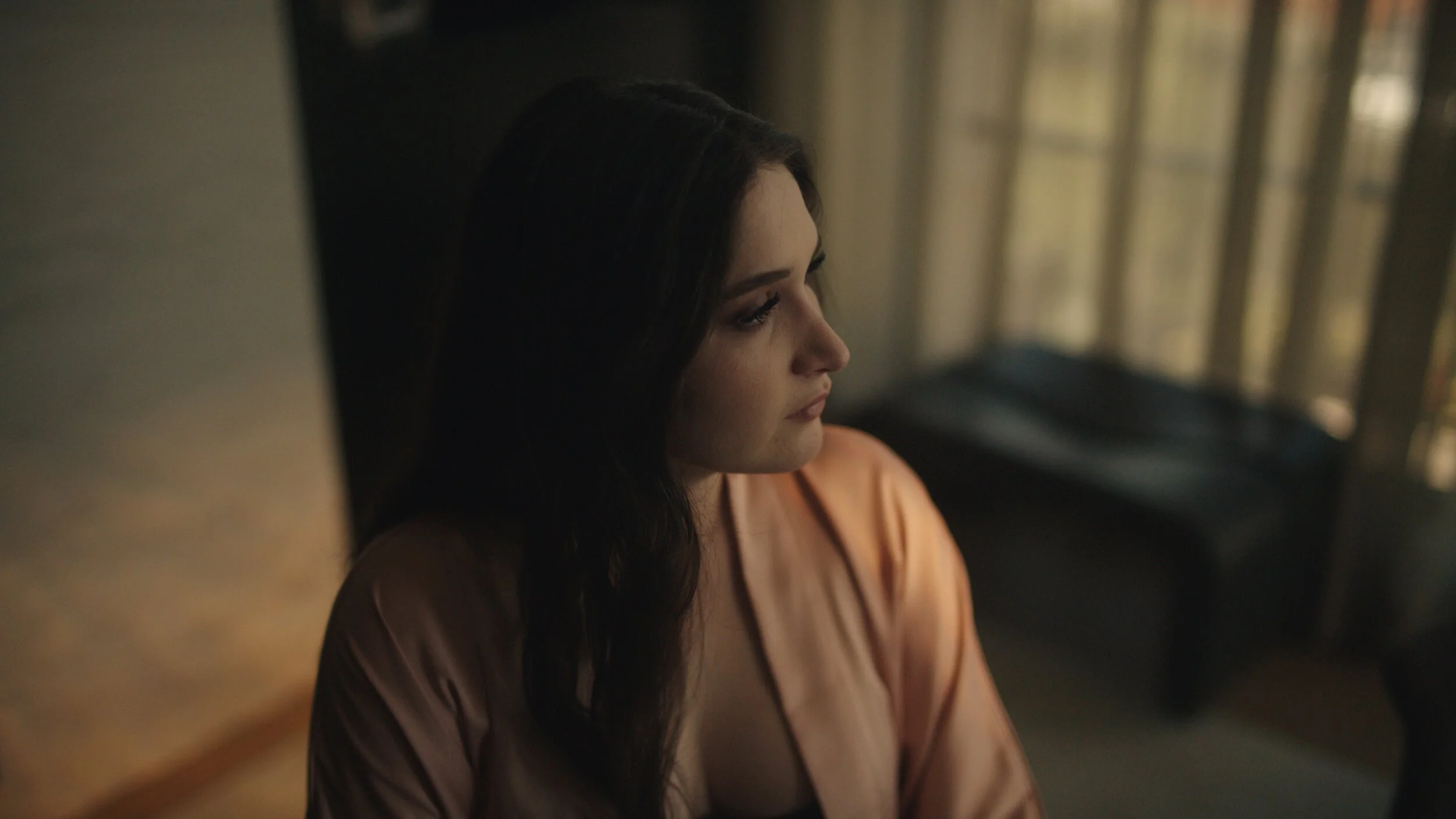 Still from the short film, FOR GOOD. In a softly lit bedroom, a woman with long dark hair stares thoughtfully out a window.