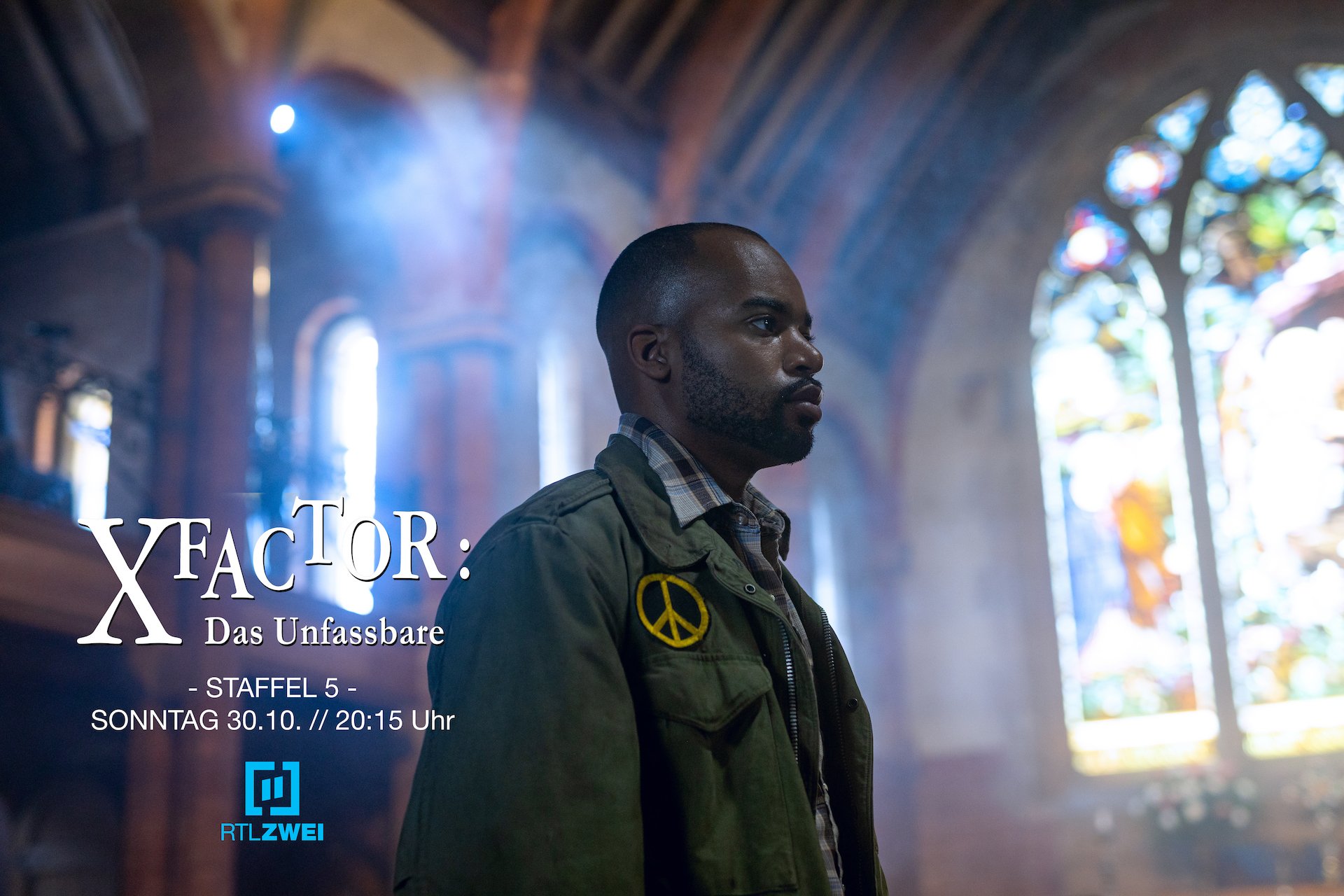 Behind-the-scenes promotional photo from the TV series X FACTOR: DAS UNFASSBARE. A bearded man stands in a church with a large stained glass window in the background.
