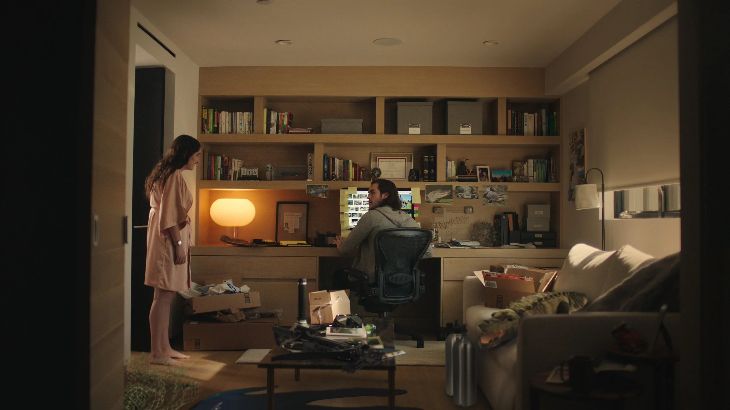 Still from the short film, FOR GOOD. A woman in a pink robe stands in a cluttered office with her husband. The room has a large bookshelf, desk lamp, and various boxes and items scattered around.