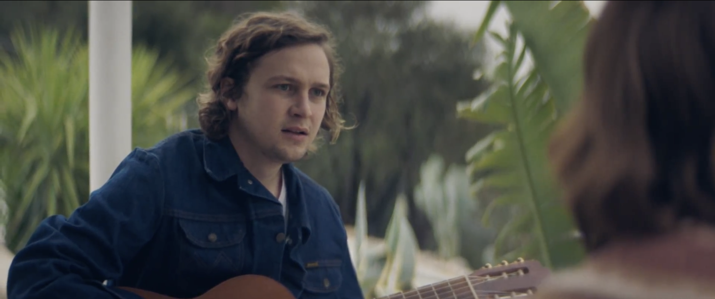 Still from the short film, SILVERTONE. A young man (Logan Miller) with curly brown hair plays an acoustic guitar while talking to a woman outside.