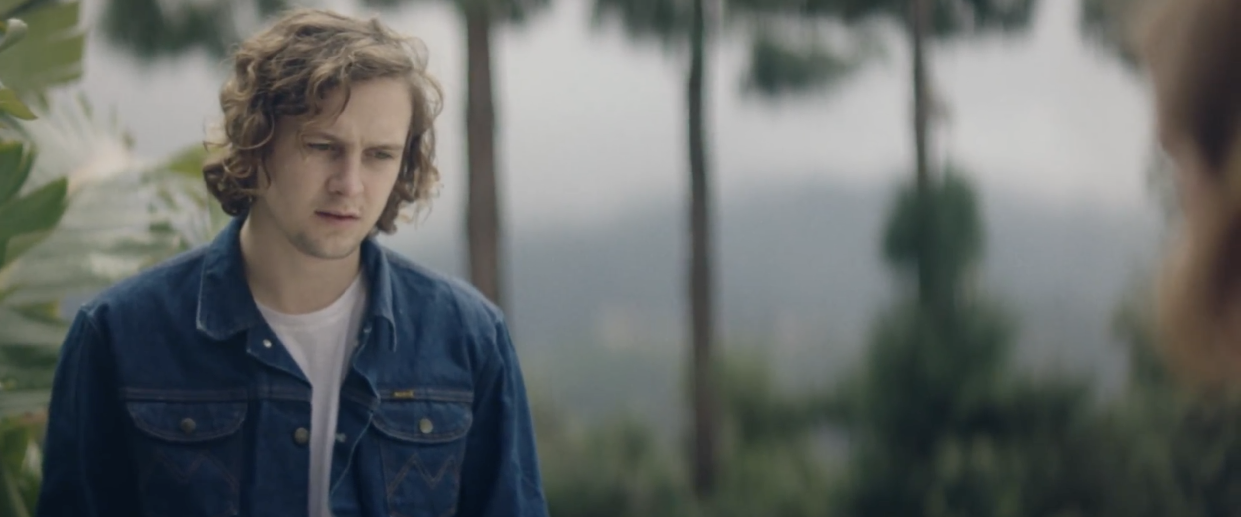 Still from the short film, SILVERTONE. A young man (Logan Miller) with curly brown hair wears a blue denim jacket. He looks thoughtful while staring at a woman (Kerris Dorsey) outside.