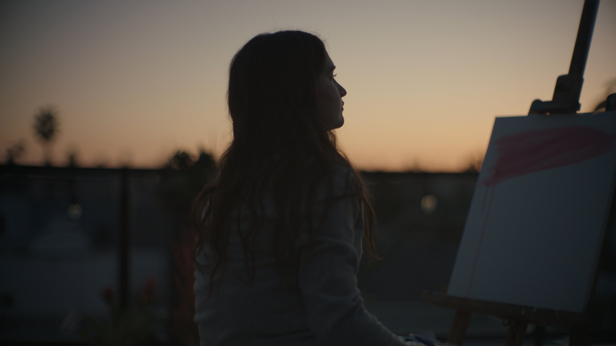 Still from the short film, FOR GOOD. A woman paints outdoors at sunset, sitting next to an easel with a canvas.