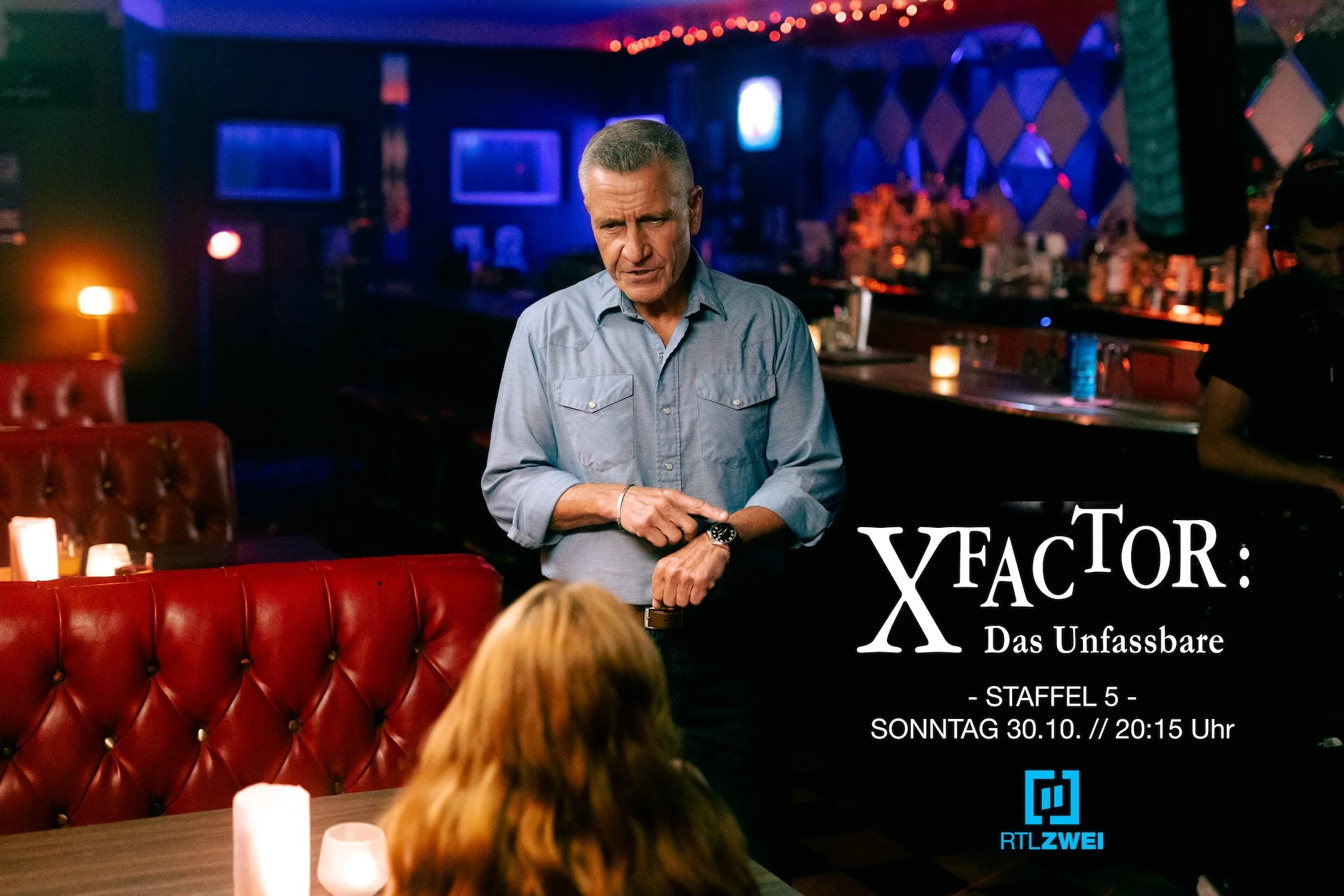 Behind-the-scenes promotional photo from the TV series X FACTOR: DAS UNFASSBARE. A man with gray hair wearing a blue button-up shirt stands in a dimly lit bar with red leather booths. He talks to a woman with red hair. 