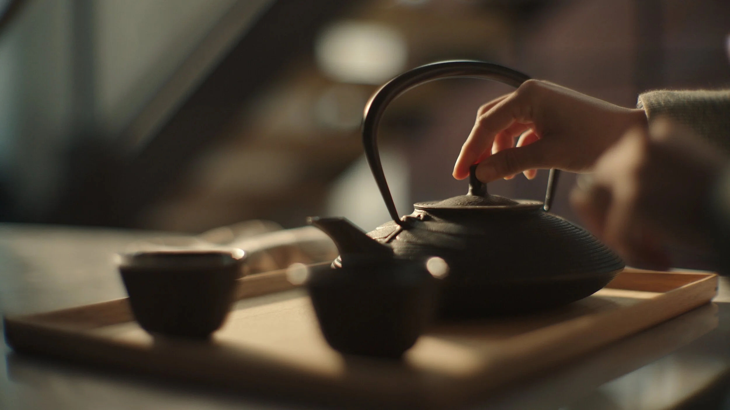 Still from the short film, FOR GOOD. A person pours tea from a teapot into a cup on a wooden tray in warm lighting.