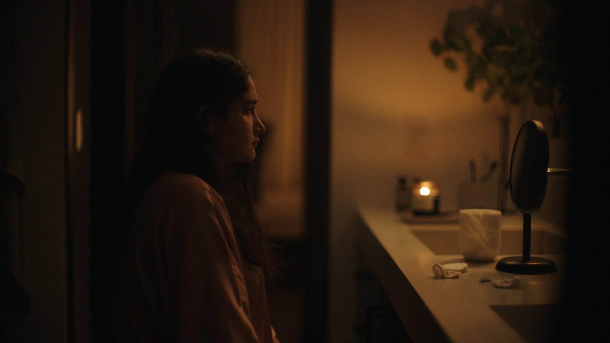 Still from the short film, FOR GOOD. A woman sits alone in a dimly lit room, facing a mirror on a countertop. The soft, warm lighting creates a contemplative atmosphere.